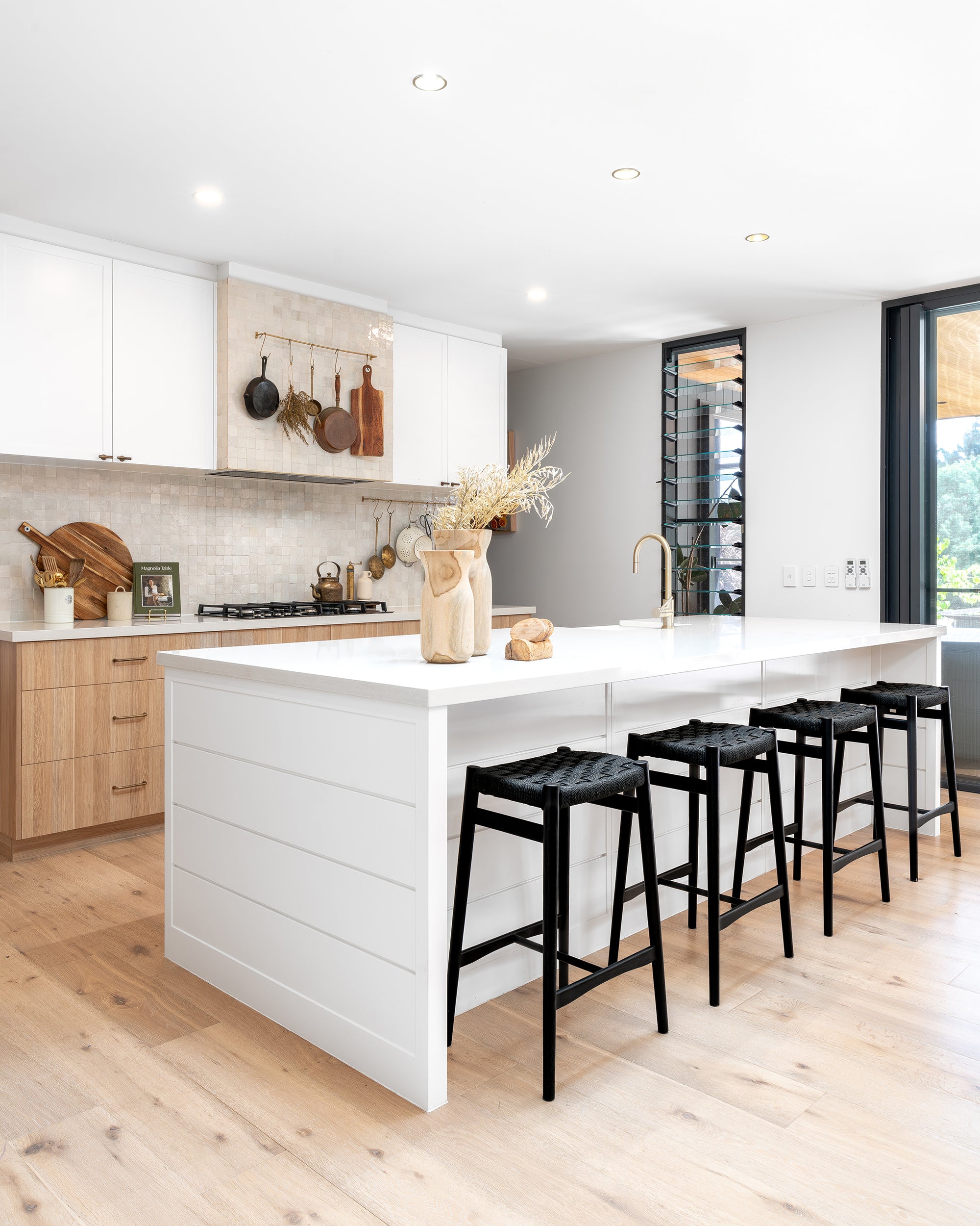 A bright and modern coastal kitchen featuring a white island with four black woven bar stools, a gold kitchen tap, and warm wooden cabinetry. The space is styled with natural elements, including wooden chopping boards, dried botanicals, and ceramic decor, creating a harmonious blend of contemporary and rustic aesthetics