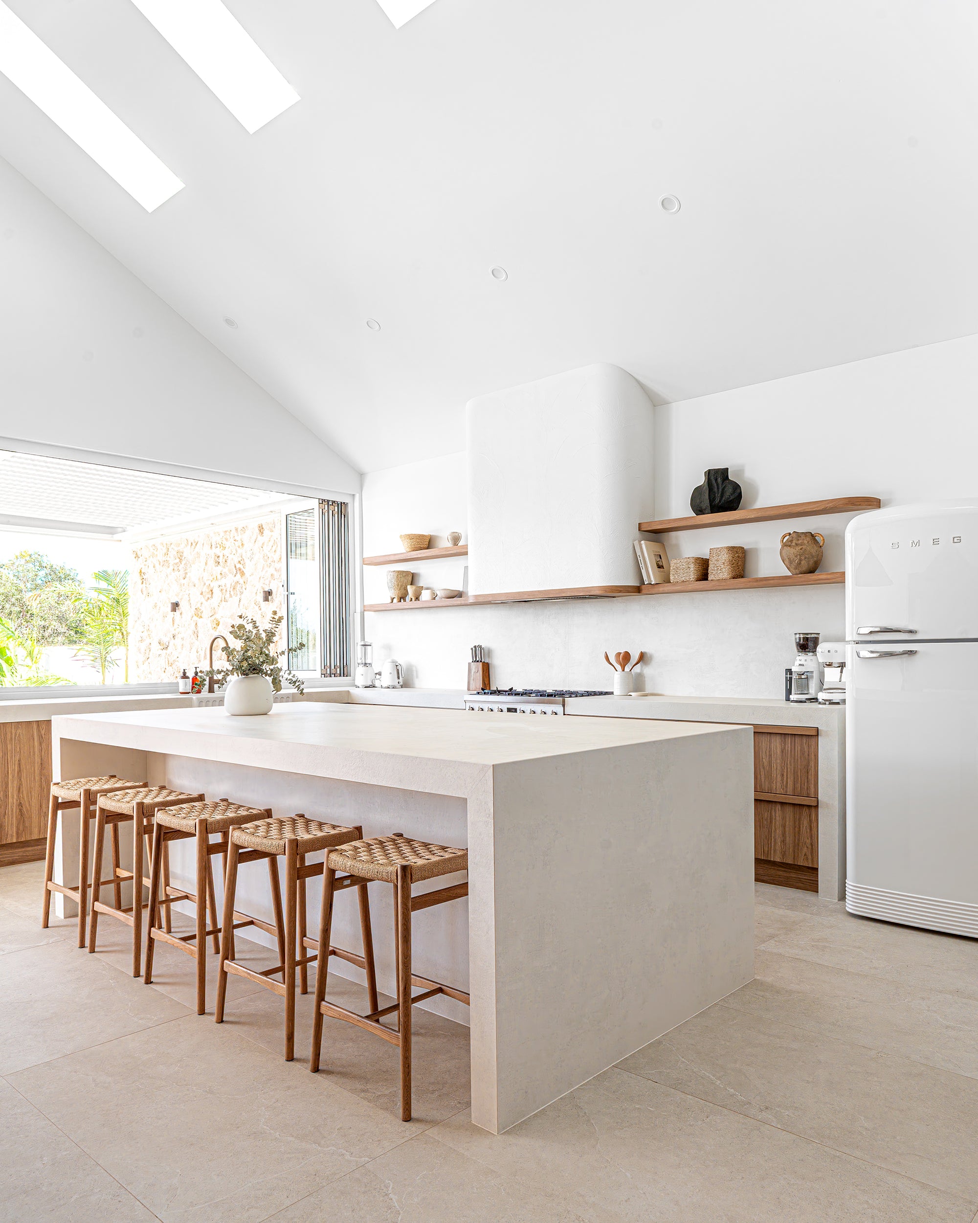 Modern coastal kitchen with a minimalist stone island, woven timber bar stools, open timber shelving, and natural light filtering through skylights.