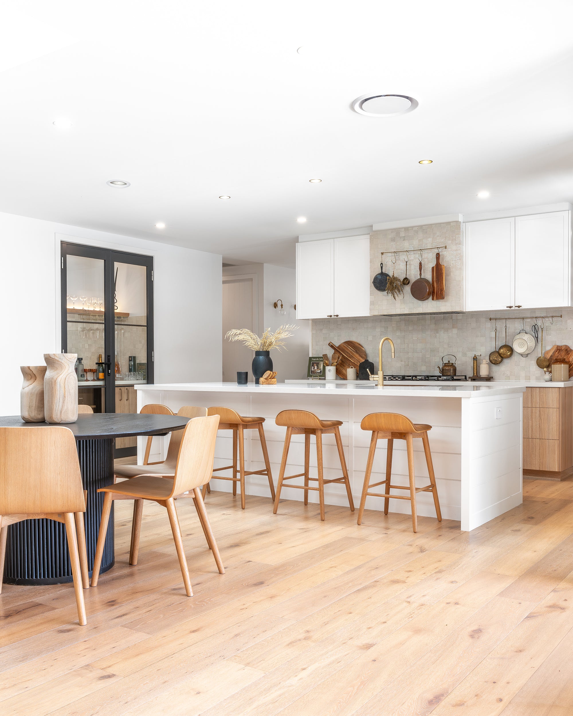 A beautifully styled modern coastal kitchen featuring a spacious island with wooden bar stools, natural timber flooring, white cabinetry, and a black dining table with matching wooden chairs, creating a warm and inviting space