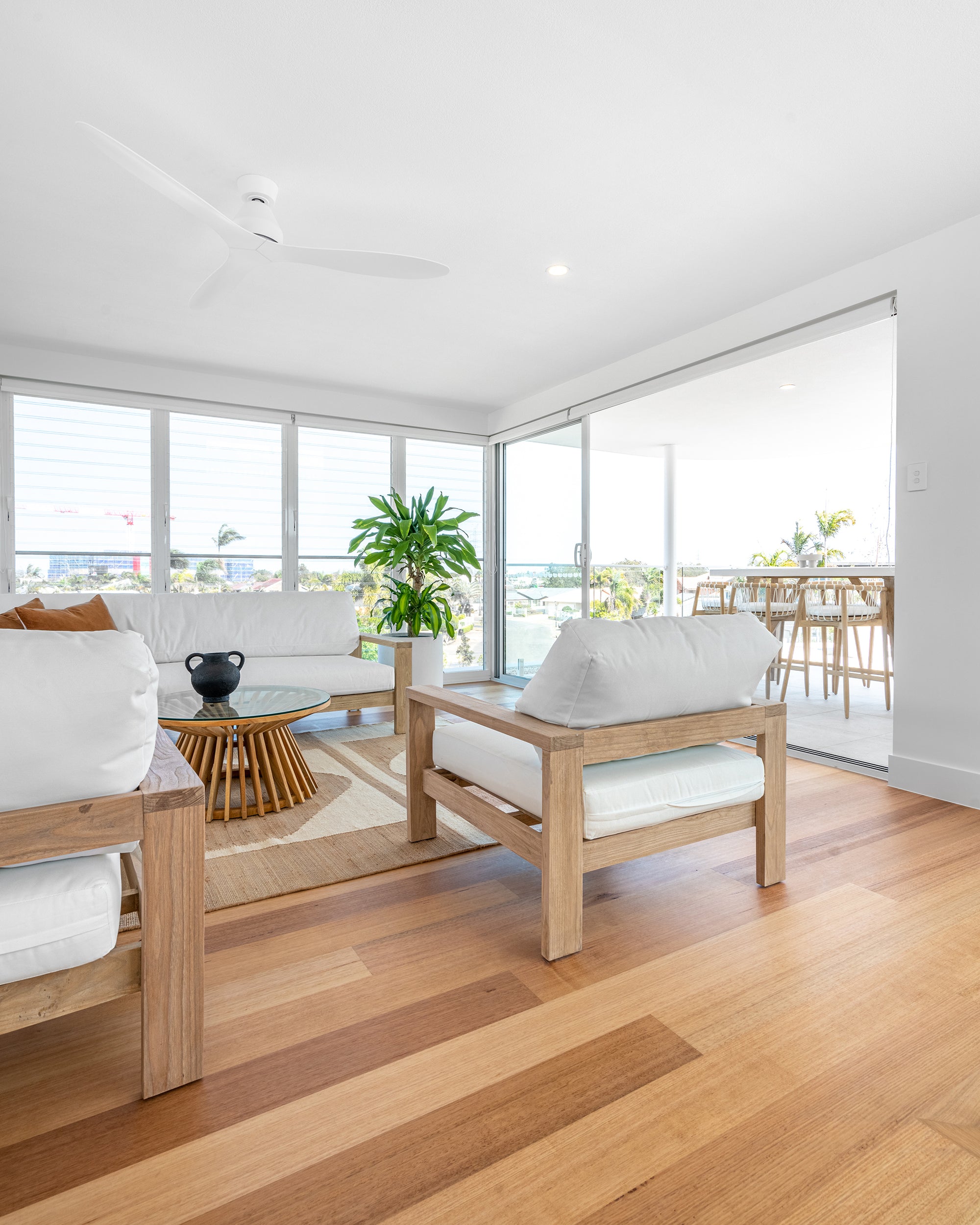 A contemporary coastal lounge setting featuring a wooden-framed sofa and armchairs with crisp white cushions, complemented by a sculptural glass-top coffee table and natural textures in a bright, open-plan space