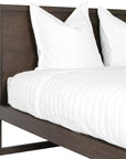 A sleek modern bed frame crafted from dark oak, featuring a minimalist open-base design. The bed is styled with crisp white linen and plush pillows, offering a refined and contemporary bedroom aesthetic