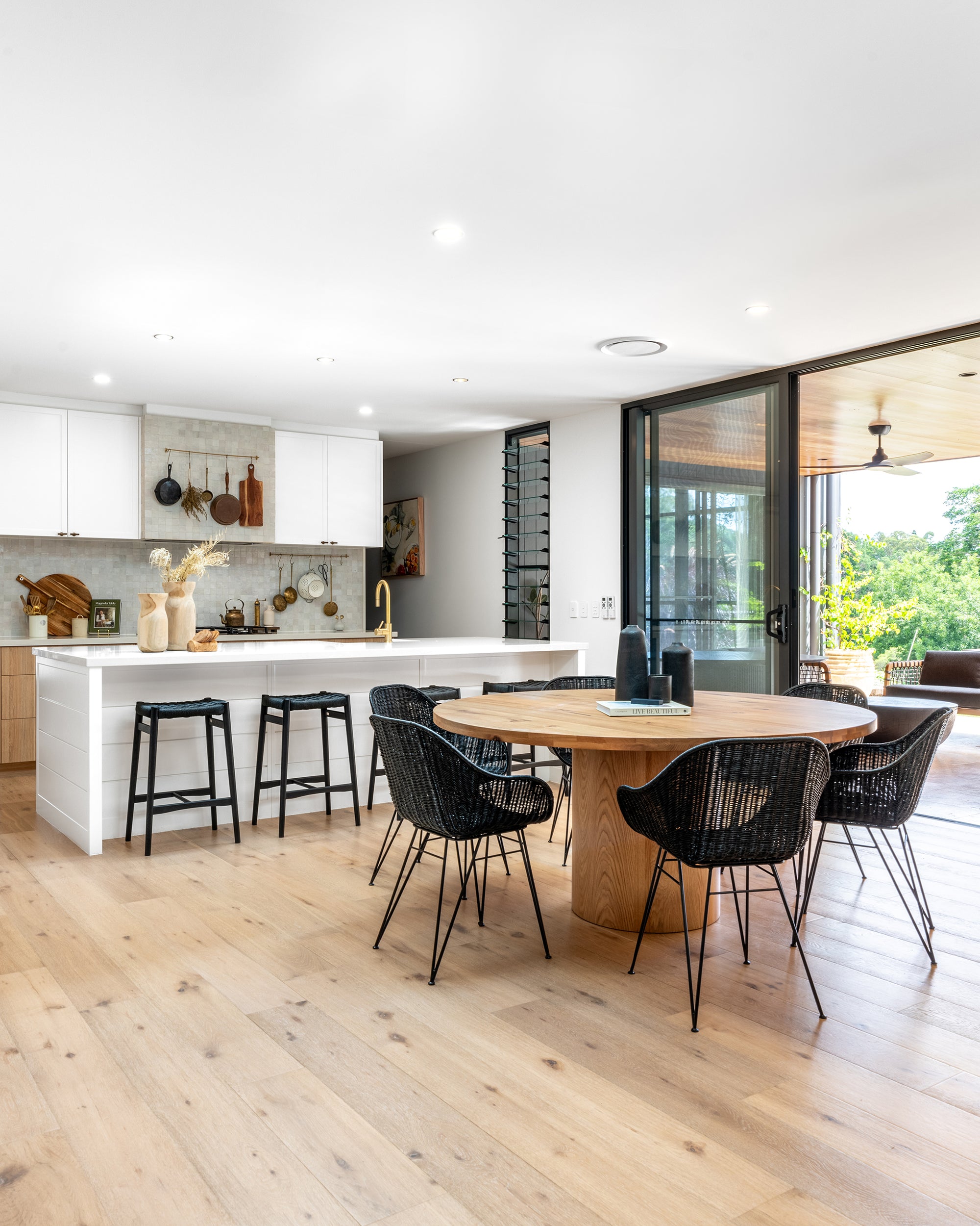 A contemporary open-plan kitchen and dining area featuring a round wooden dining table surrounded by black woven chairs, paired with a white kitchen island with matching black bar stools. The space is styled with natural wood tones, brass accents, and ceramic decor, creating a warm and inviting modern aesthetic with a seamless indoor-outdoor flow