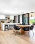 A contemporary open-plan kitchen and dining area featuring a round wooden dining table surrounded by black woven chairs, paired with a white kitchen island with matching black bar stools. The space is styled with natural wood tones, brass accents, and ceramic decor, creating a warm and inviting modern aesthetic with a seamless indoor-outdoor flow