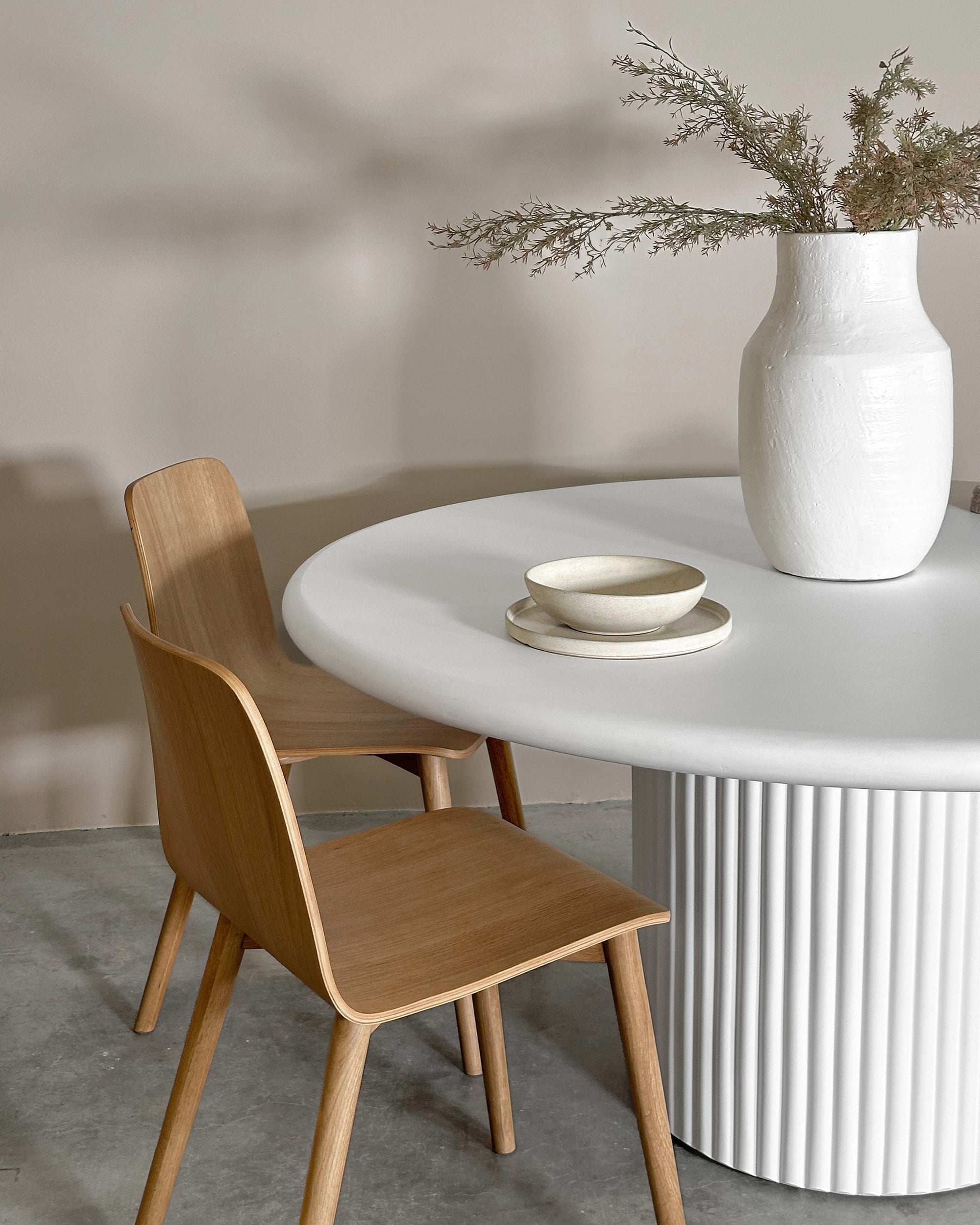 A modern dining space featuring a white round table with a fluted base, paired with minimalist wooden chairs. A neutral ceramic bowl set and a textured white vase with dried foliage add organic elegance to the setting