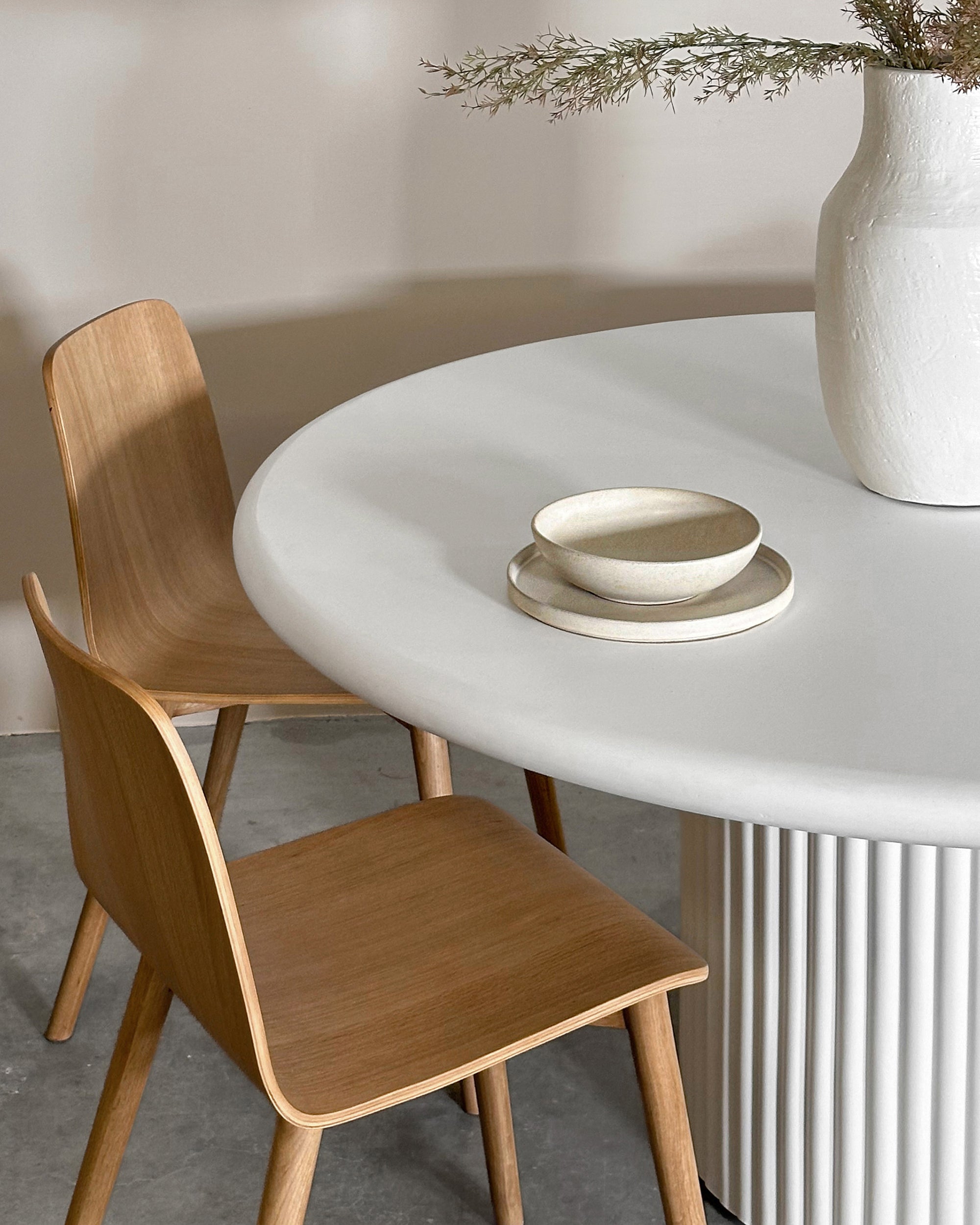 A modern dining space featuring a white round table with a fluted base, paired with minimalist wooden chairs. A neutral ceramic bowl set and a textured white vase with dried foliage add organic elegance to the setting