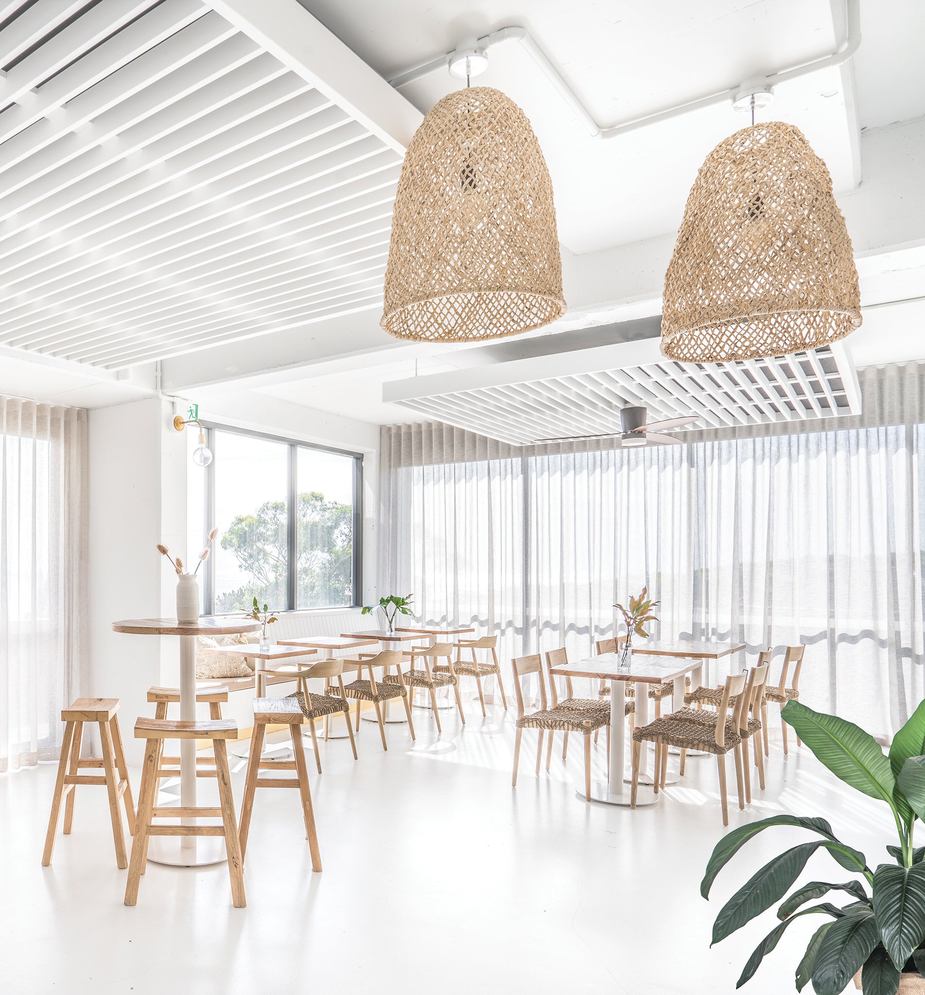 Bright and airy modern dining space with woven natural fibre pendant lights, wooden dining chairs with woven seats, and tall bar stools around round wooden tables.