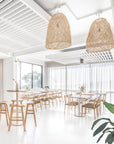 Bright and airy modern dining space with woven natural fibre pendant lights, wooden dining chairs with woven seats, and tall bar stools around round wooden tables.