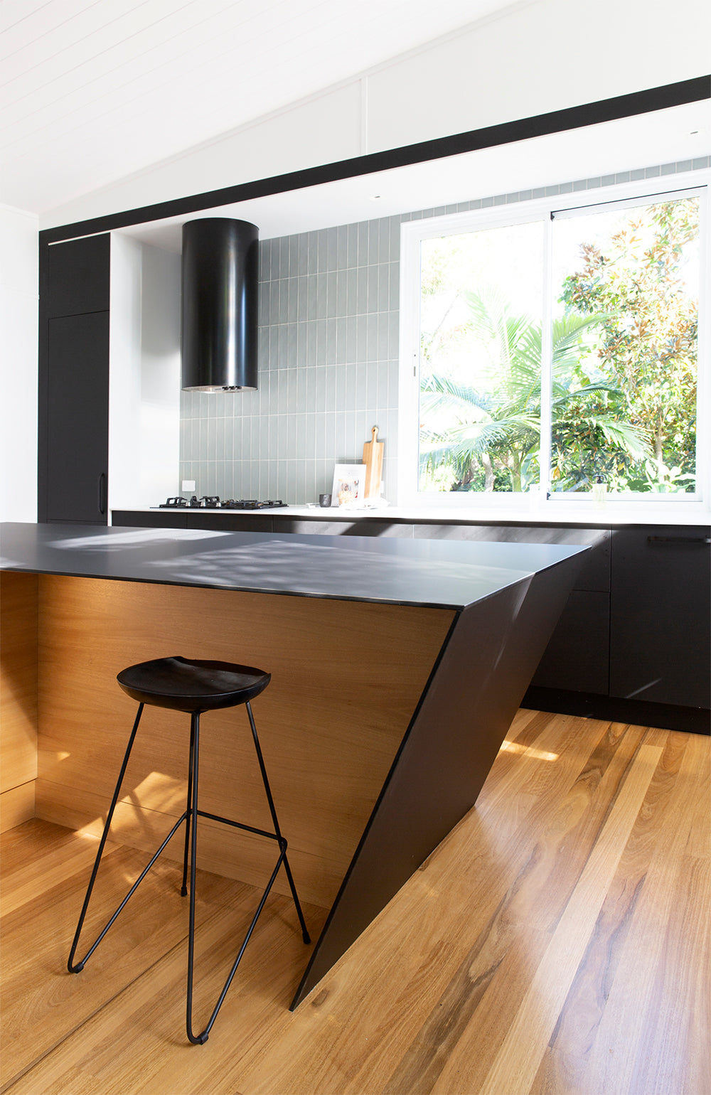 A contemporary kitchen featuring Kulala Living black bar stools with a minimalist metal frame, paired with a sleek wooden island. The space is accented by bold black cabinetry, soft grey tiles, and a striking gold statement pendant light
