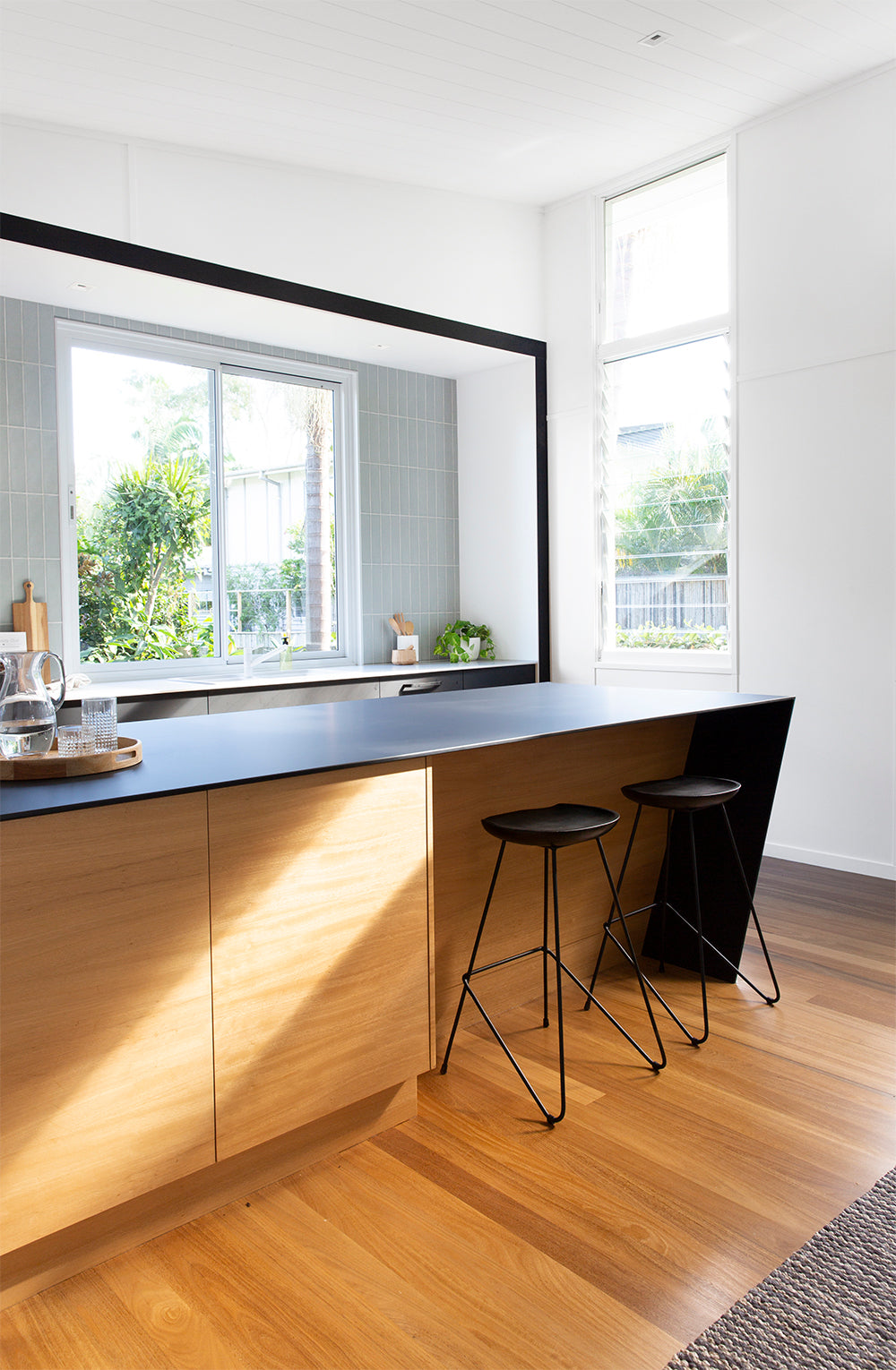 A contemporary kitchen featuring Kulala Living black bar stools with a minimalist metal frame, paired with a sleek wooden island. The space is accented by bold black cabinetry, soft grey tiles, and a striking gold statement pendant light