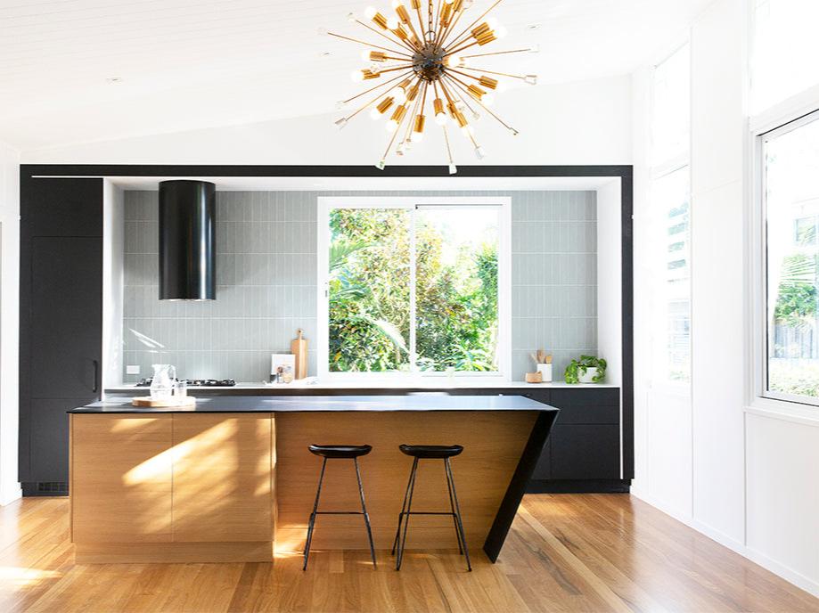 A contemporary kitchen featuring Kulala Living black bar stools with a minimalist metal frame, paired with a sleek wooden island. The space is accented by bold black cabinetry, soft grey tiles, and a striking gold statement pendant light