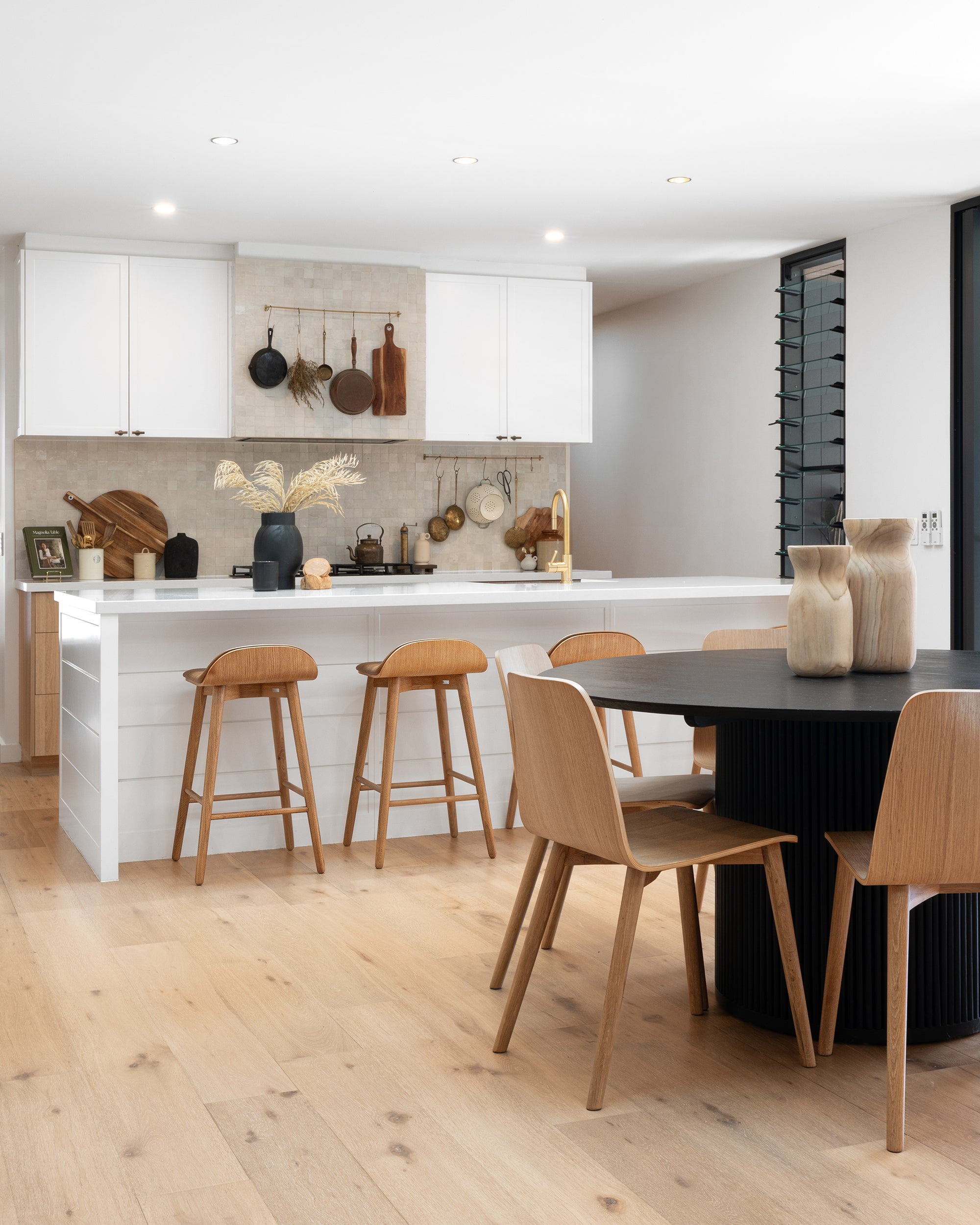A contemporary kitchen featuring a sleek white island with natural wood bar stools, a black round dining table, and oak dining chairs. The space is styled with organic textures, minimalist cabinetry, and warm neutral tones, creating a relaxed and inviting atmosphere