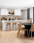 A contemporary kitchen featuring a sleek white island with natural wood bar stools, a black round dining table, and oak dining chairs. The space is styled with organic textures, minimalist cabinetry, and warm neutral tones, creating a relaxed and inviting atmosphere