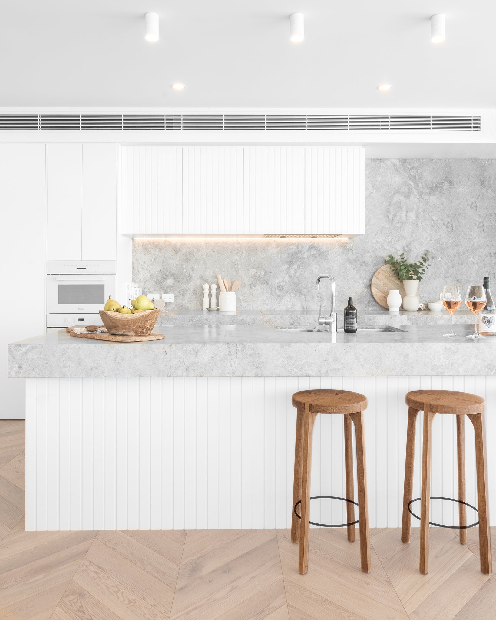 A bright modern kitchen featuring Kulala Living solid oak bar stools with black metal footrests. Styled with a marble benchtop, white cabinetry, and natural wood accents for a contemporary coastal aesthetic