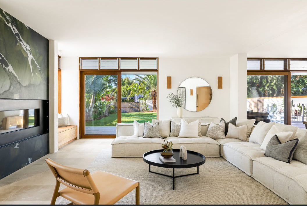 A stylish modern lounge featuring a large white linen sectional sofa adorned with neutral-toned cushions, a round black coffee table, and a leather accent chair. The space is illuminated by natural light from large glass doors, showcasing lush greenery outside