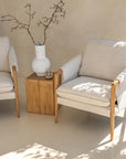 Kulala Living modern lounge setting featuring two neutral upholstered armchairs with wooden armrests, paired with a block side table and a white textured vase with branches. A serene and stylish outdoor retreat