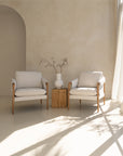 Kulala Living modern lounge setting featuring two neutral upholstered armchairs with wooden armrests, paired with a block side table and a white textured vase with branches. A serene and stylish outdoor retreat