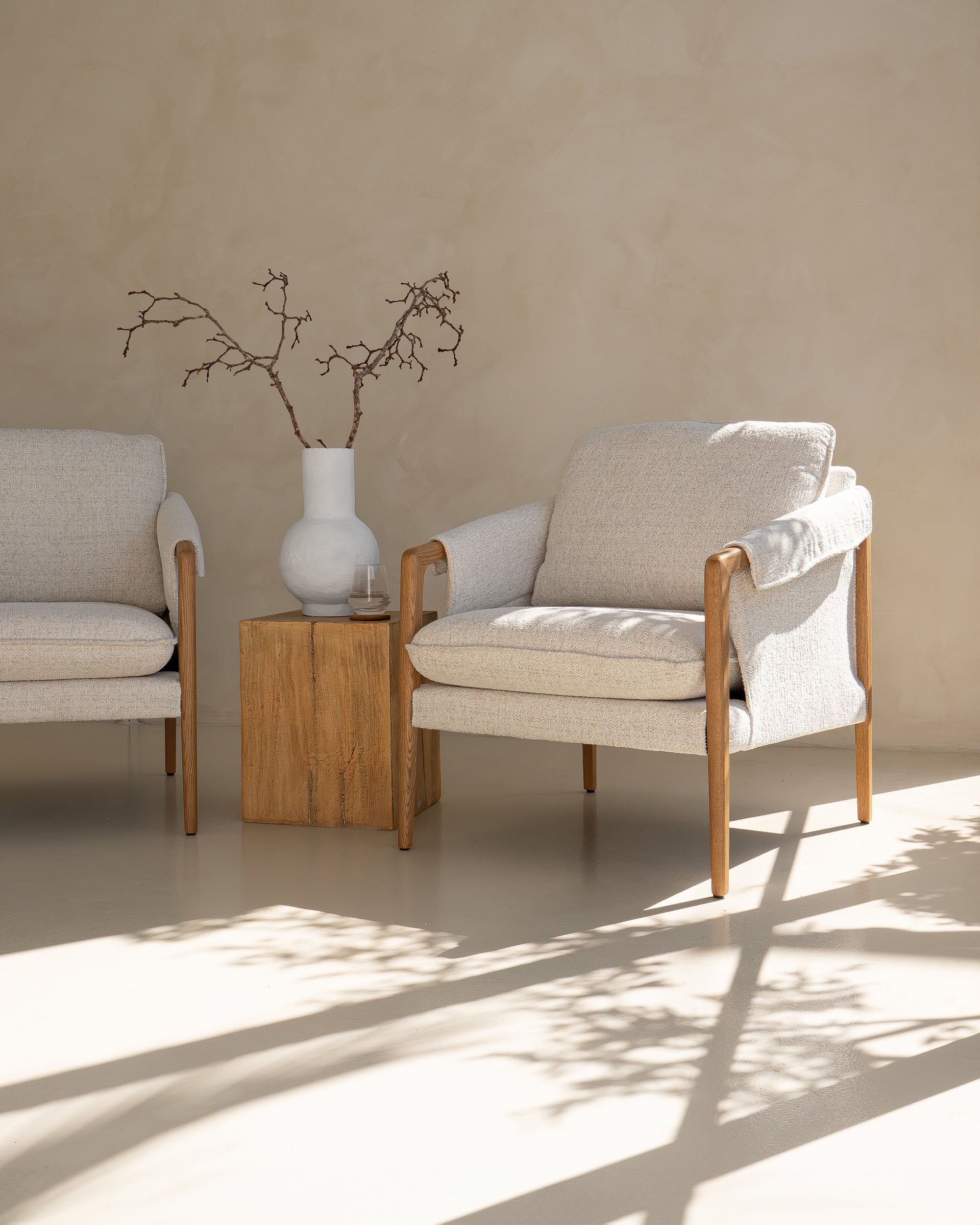 Kulala Living modern lounge setting featuring two neutral upholstered armchairs with wooden armrests, paired with a block side table and a white textured vase with branches. A serene and stylish outdoor retreat