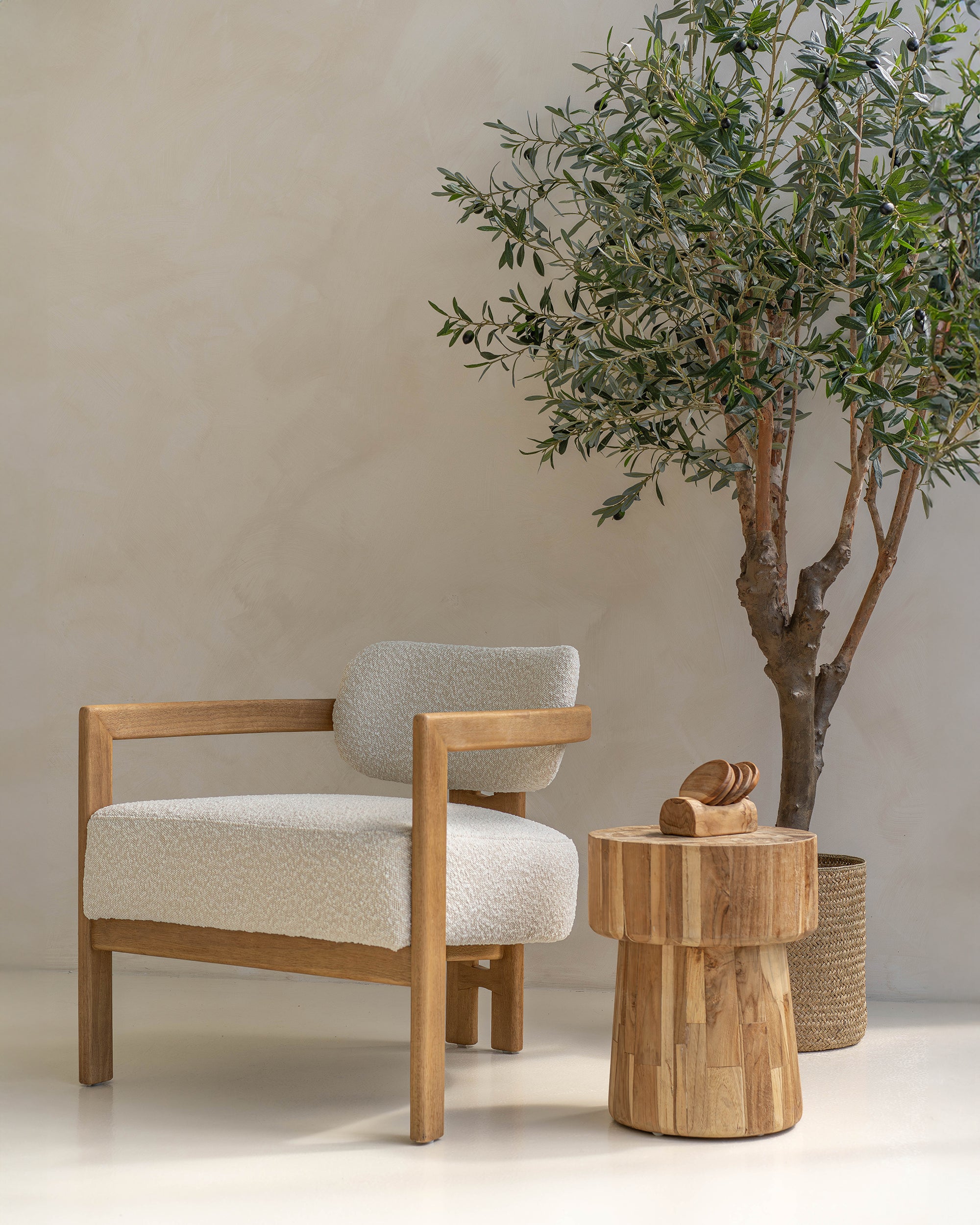 Contemporary wooden armchair with boucle upholstery, paired with a rustic timber side table and an olive tree in a woven basket, set against a neutral-toned plaster wall