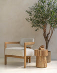 Contemporary wooden armchair with boucle upholstery, paired with a rustic timber side table and an olive tree in a woven basket, set against a neutral-toned plaster wall