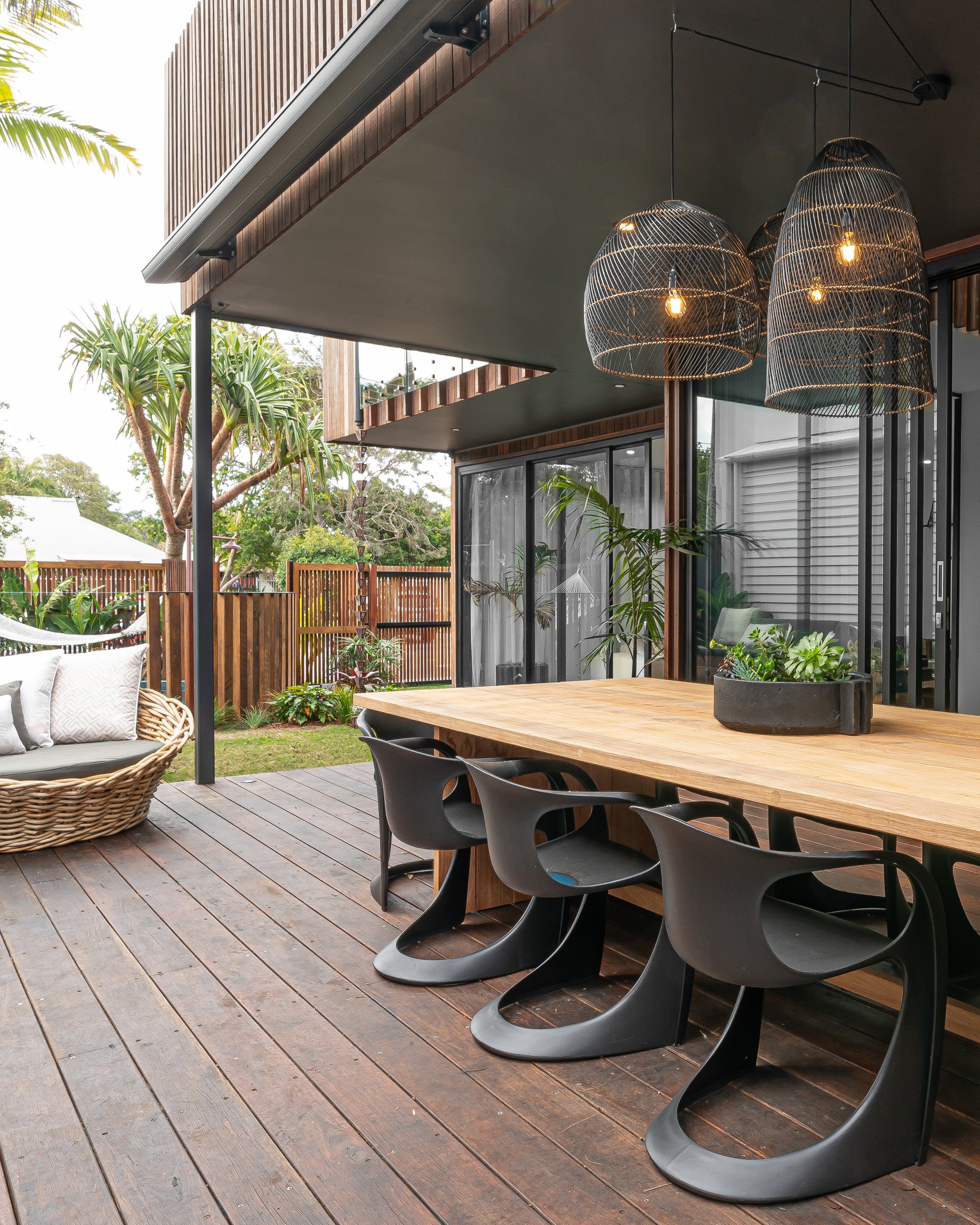 Modern outdoor dining area with black woven pendant lights, timber table, sleek black chairs, and natural landscaping