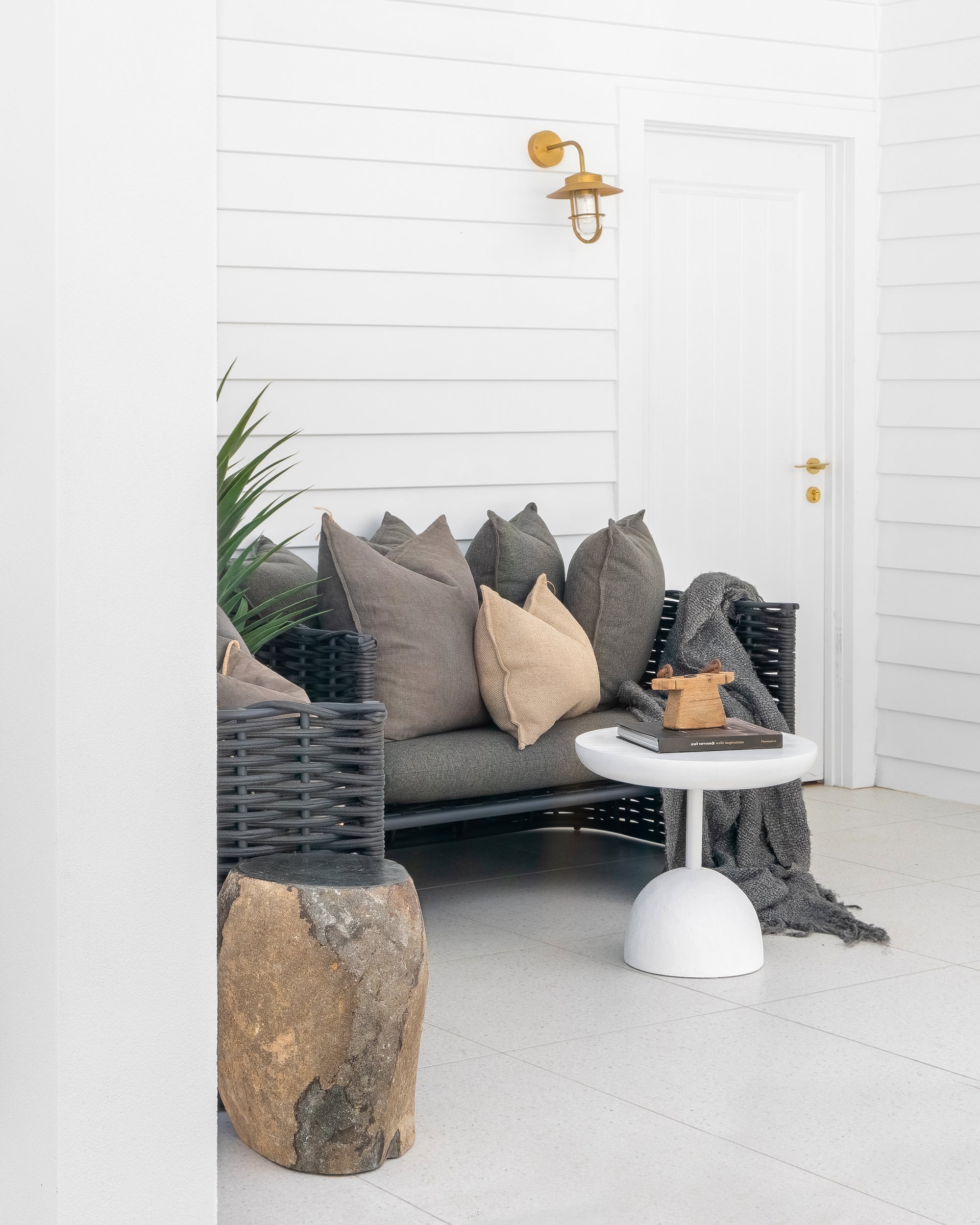 A stylish outdoor lounge setting featuring a black woven sofa with neutral-toned cushions, a white sculptural side table, and a natural stone stool, creating a sophisticated and relaxed atmosphere.