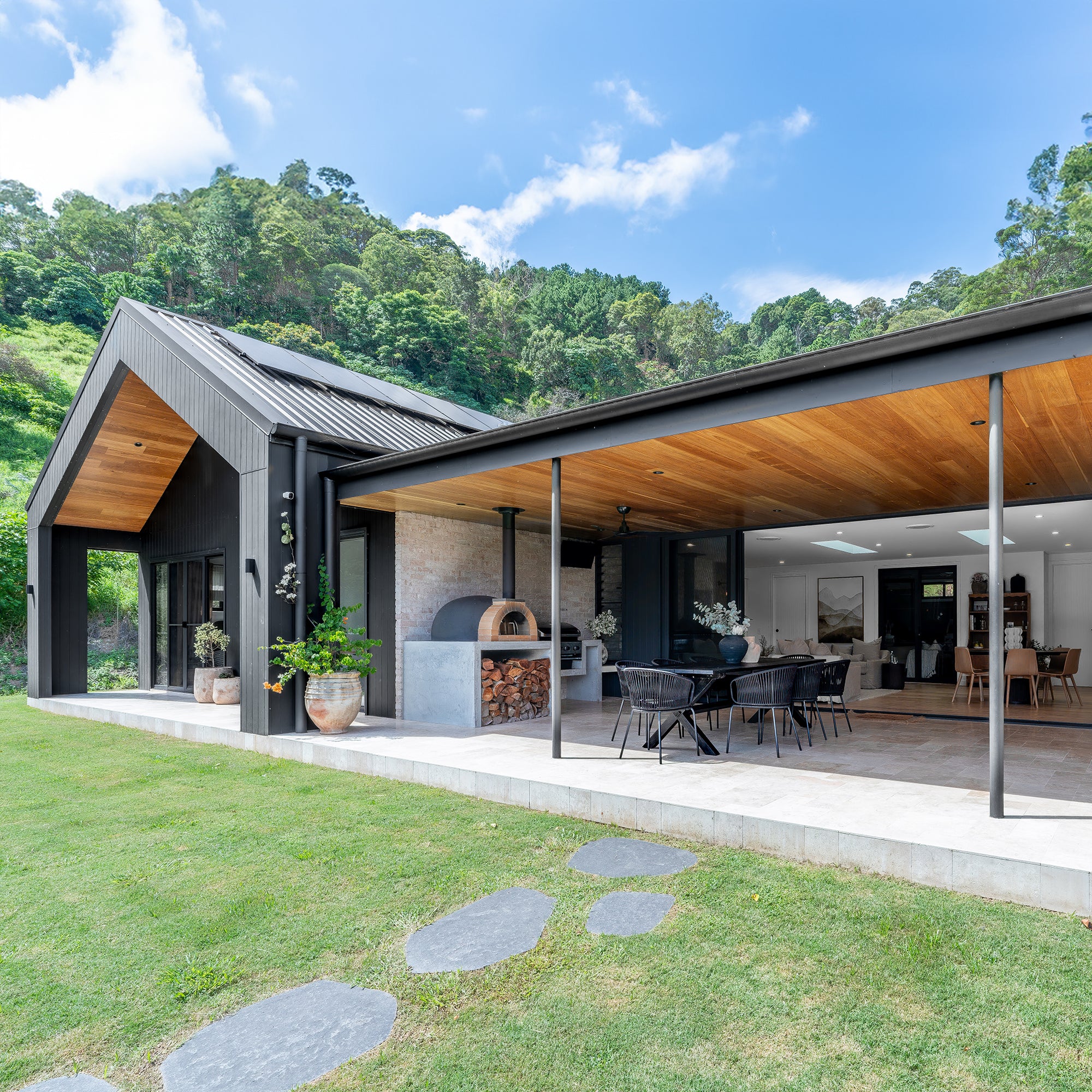 A stylish outdoor dining space featuring a black gable house with timber ceiling details, an open-plan living area, and a built-in wood-fired pizza oven. Surrounded by lush greenery, this modern retreat blends contemporary architecture with natural elements