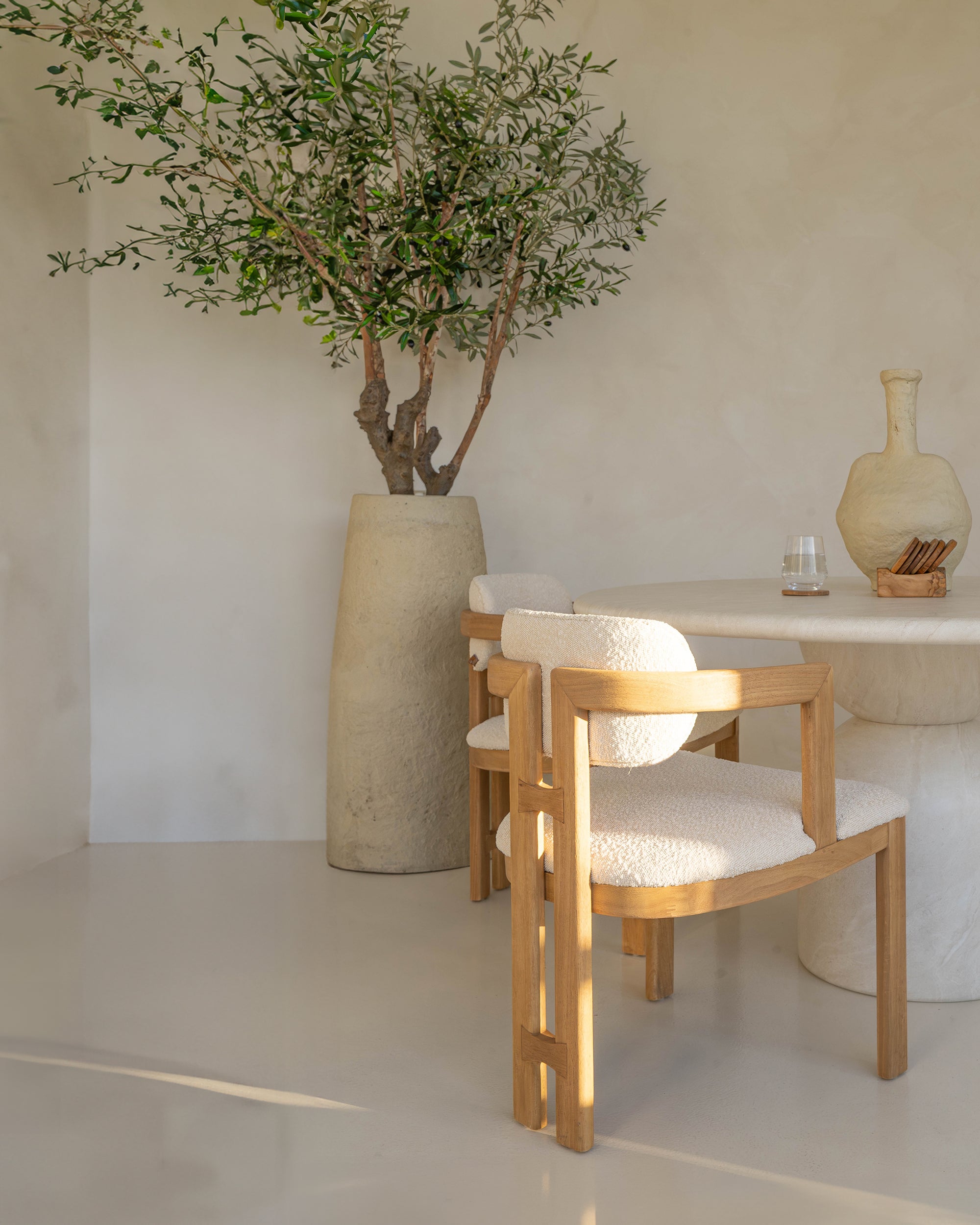 Stylish wooden dining chair with boucle upholstery, set in a minimalist dining space with a sculptural stone table and an olive tree in a textured planter.
