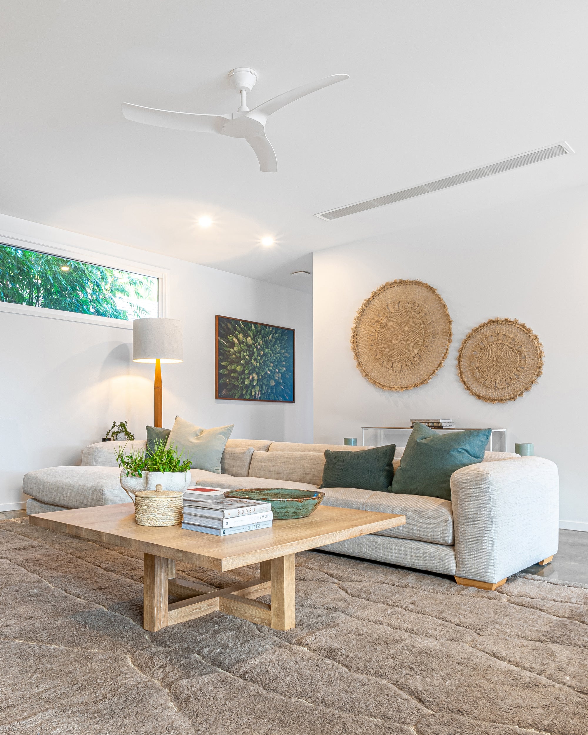 Modern lounge room with a natural aesthetic, featuring a neutral modular sofa with green cushions, a wooden coffee table with decor accents, woven wall hangings, and a textured floor rug
