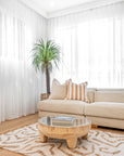 Modern lounge room featuring a neutral-toned sofa with striped cushion, wooden round coffee table with glass top, woven planter with indoor plant, and sheer white curtains