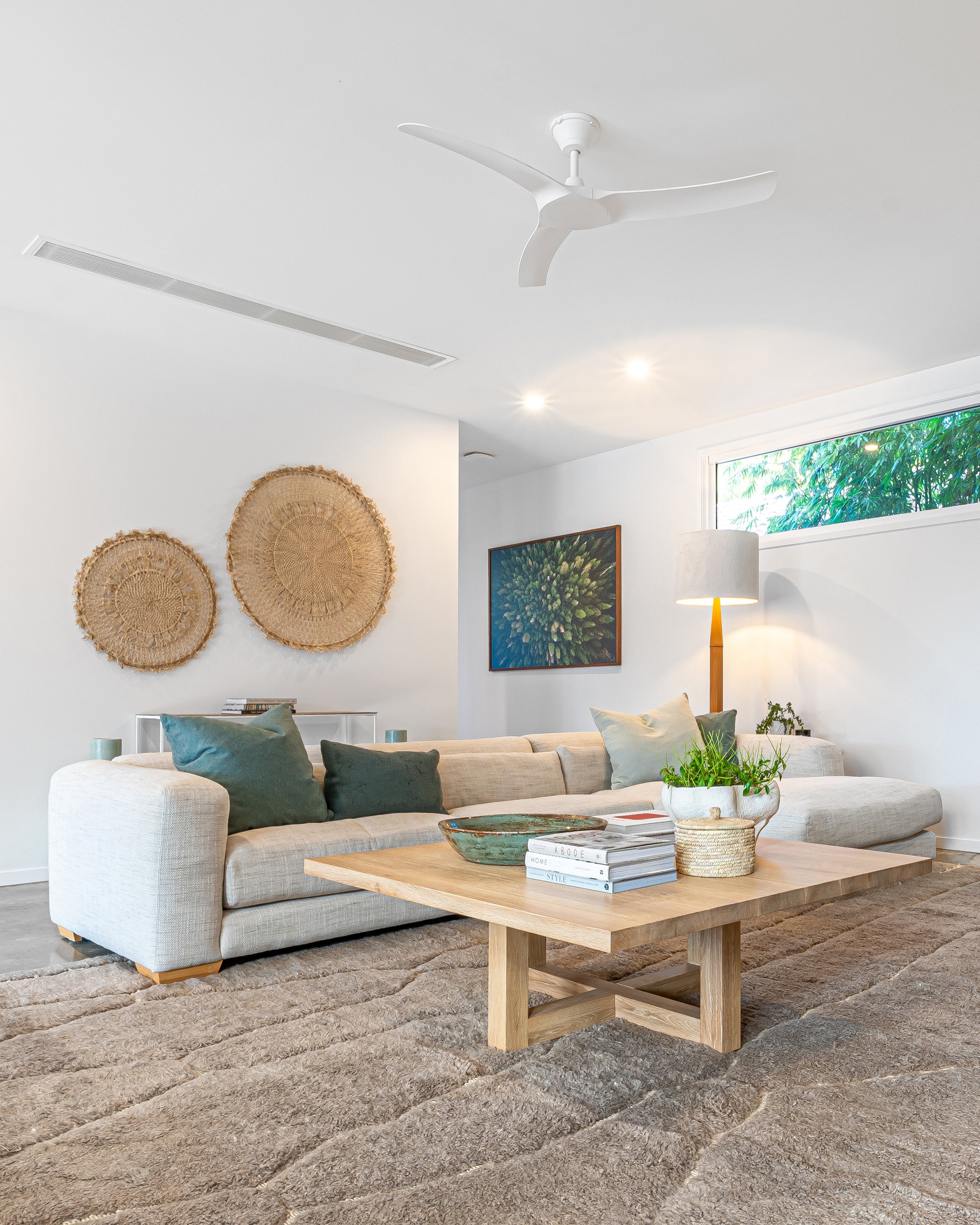 Modern lounge room with a natural aesthetic, featuring a neutral modular sofa with green cushions, a wooden coffee table with decor accents, woven wall hangings, and a textured floor rug.