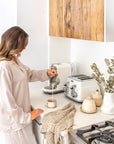 Woman pouring milk into a ceramic coffee cup in a neutral-toned kitchen with linen textures and artisan ceramics