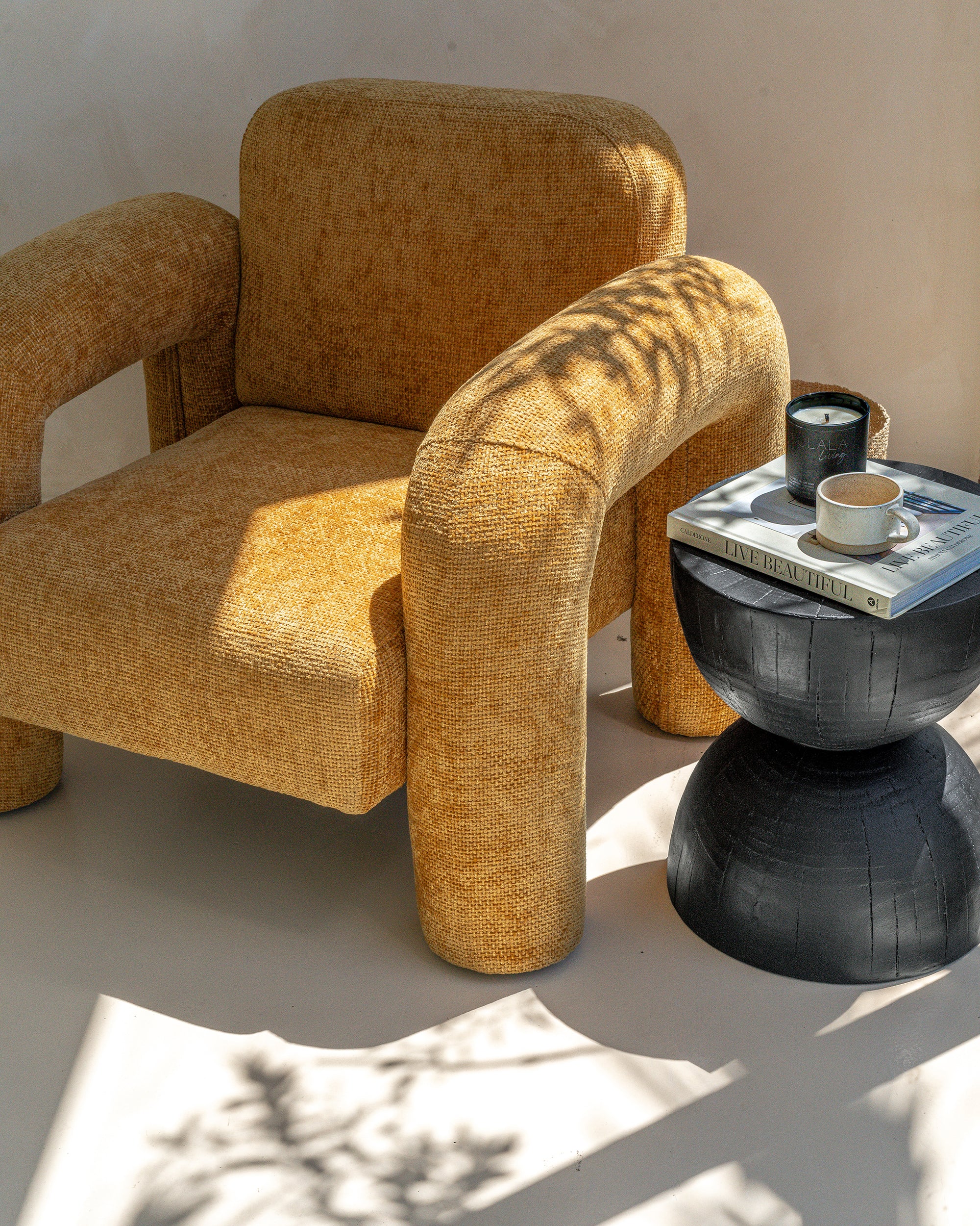A modern mustard bouclé armchair with curved lines, paired with a round black sculptural side table. Styled in a sunlit space with natural shadows and earthy tones, creating a warm and inviting setting