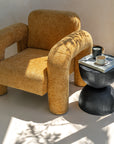A modern mustard bouclé armchair with curved lines, paired with a round black sculptural side table. Styled in a sunlit space with natural shadows and earthy tones, creating a warm and inviting setting