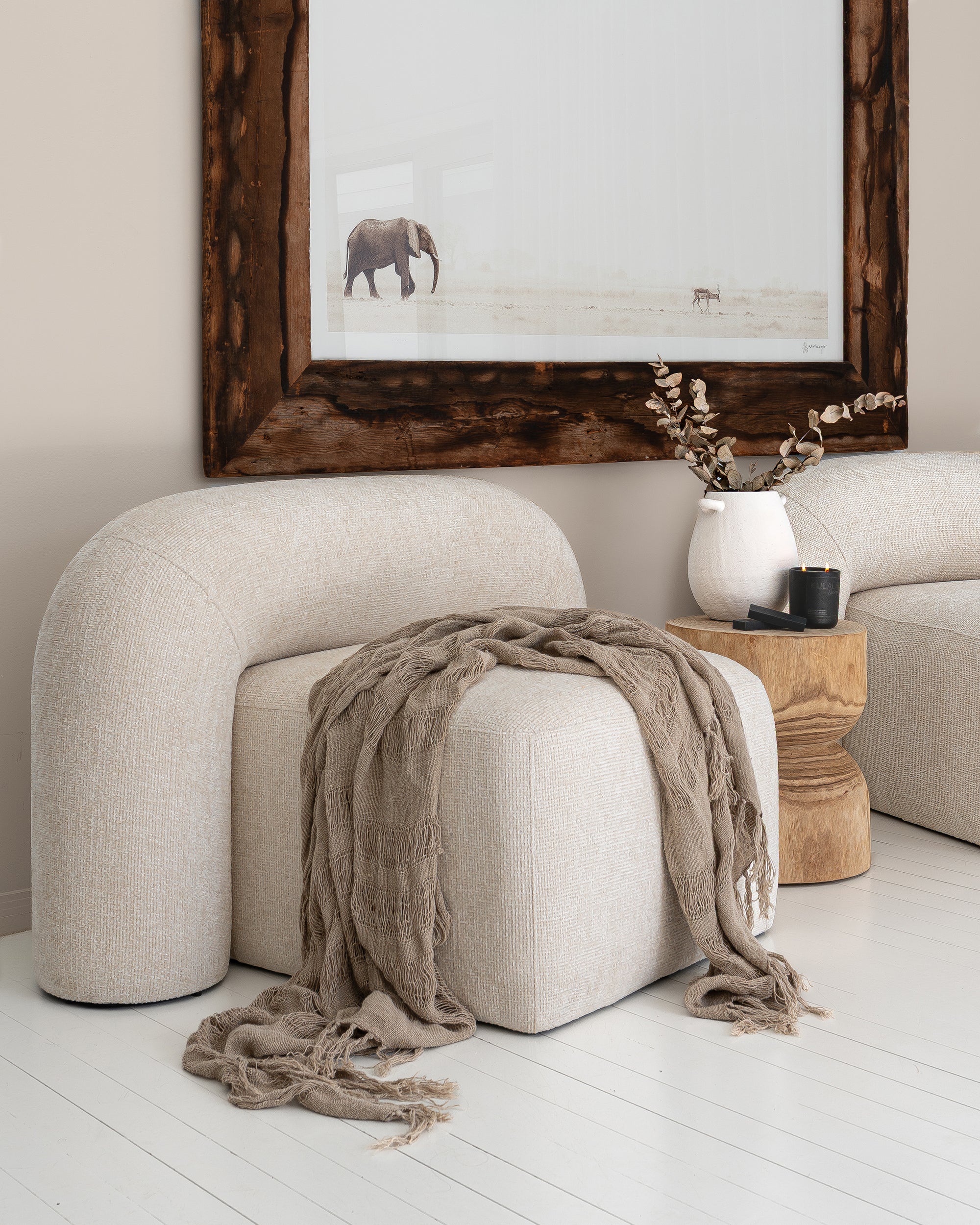 Curved boucle nash chair with a draped taupe throw, styled alongside a sculptural timber side table, ceramic vase, and candle, beneath framed elephant wall art.