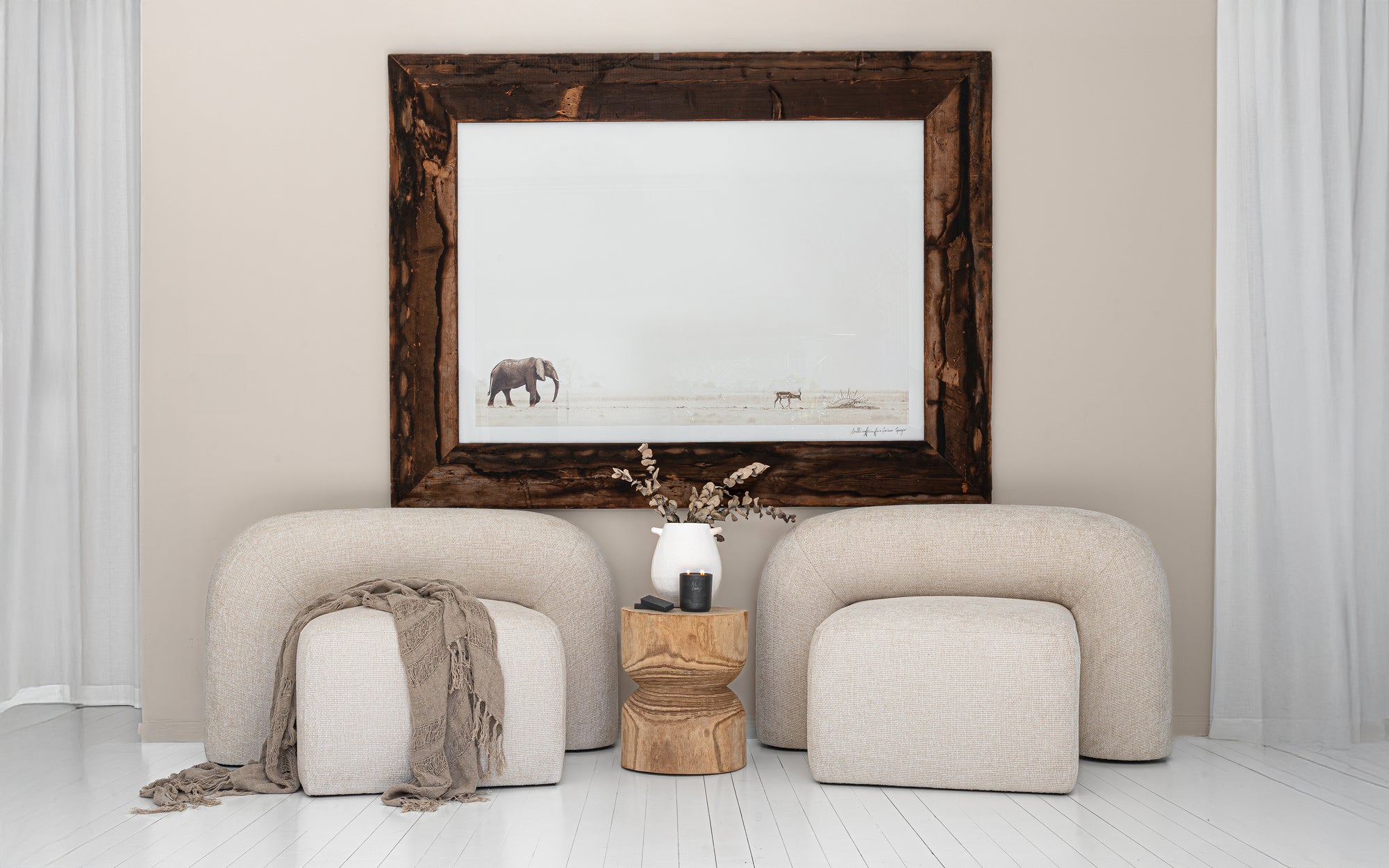 Nash chairs in natural upholstery styled with a timber side table, ceramic vase, and elephant wall art in a bright, whitewashed interior.