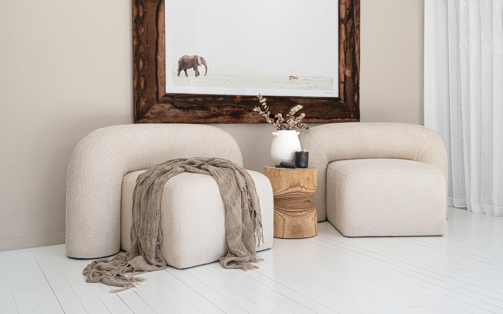 Nash occasional chairs in natural upholstery styled with a timber side table, ceramic vase, and elephant wall art in a bright, whitewashed interior.