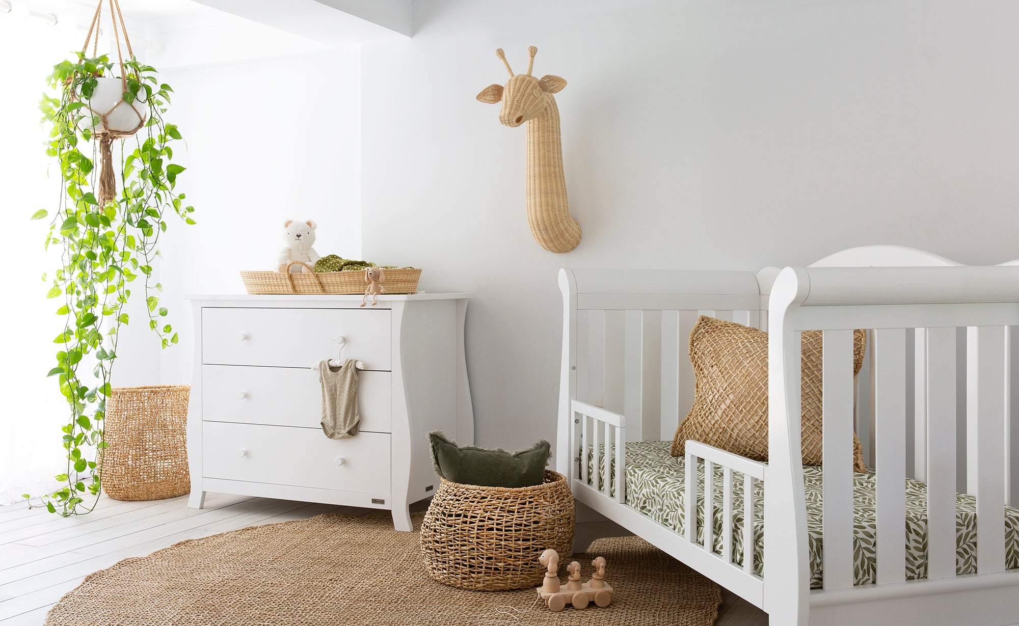 A serene nursery setting featuring woven storage baskets, a white wooden cot, a matching dresser, and a macrame hanging planter, creating a warm and natural space for a baby