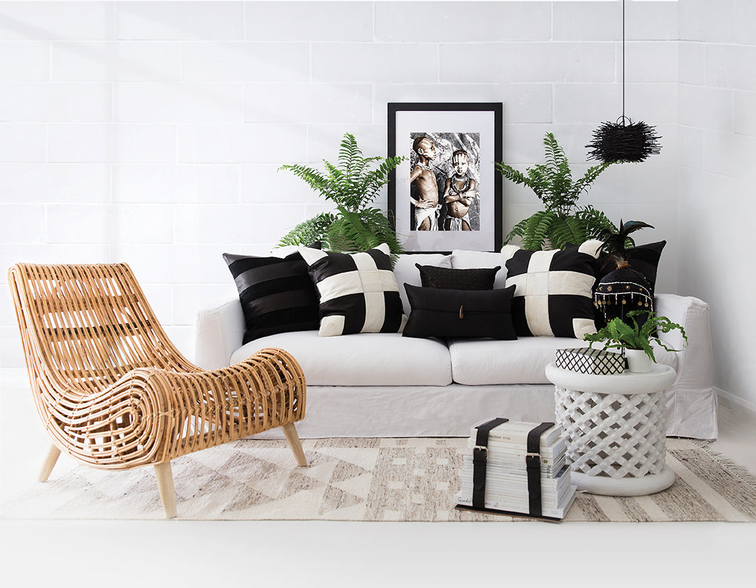 A handwoven natural rattan lounge chair with a sculptural design, styled in a contemporary living space featuring a white linen sofa, black-and-white geometric cushions, and lush green ferns. The setting blends modern and organic textures for a relaxed yet sophisticated aesthetic