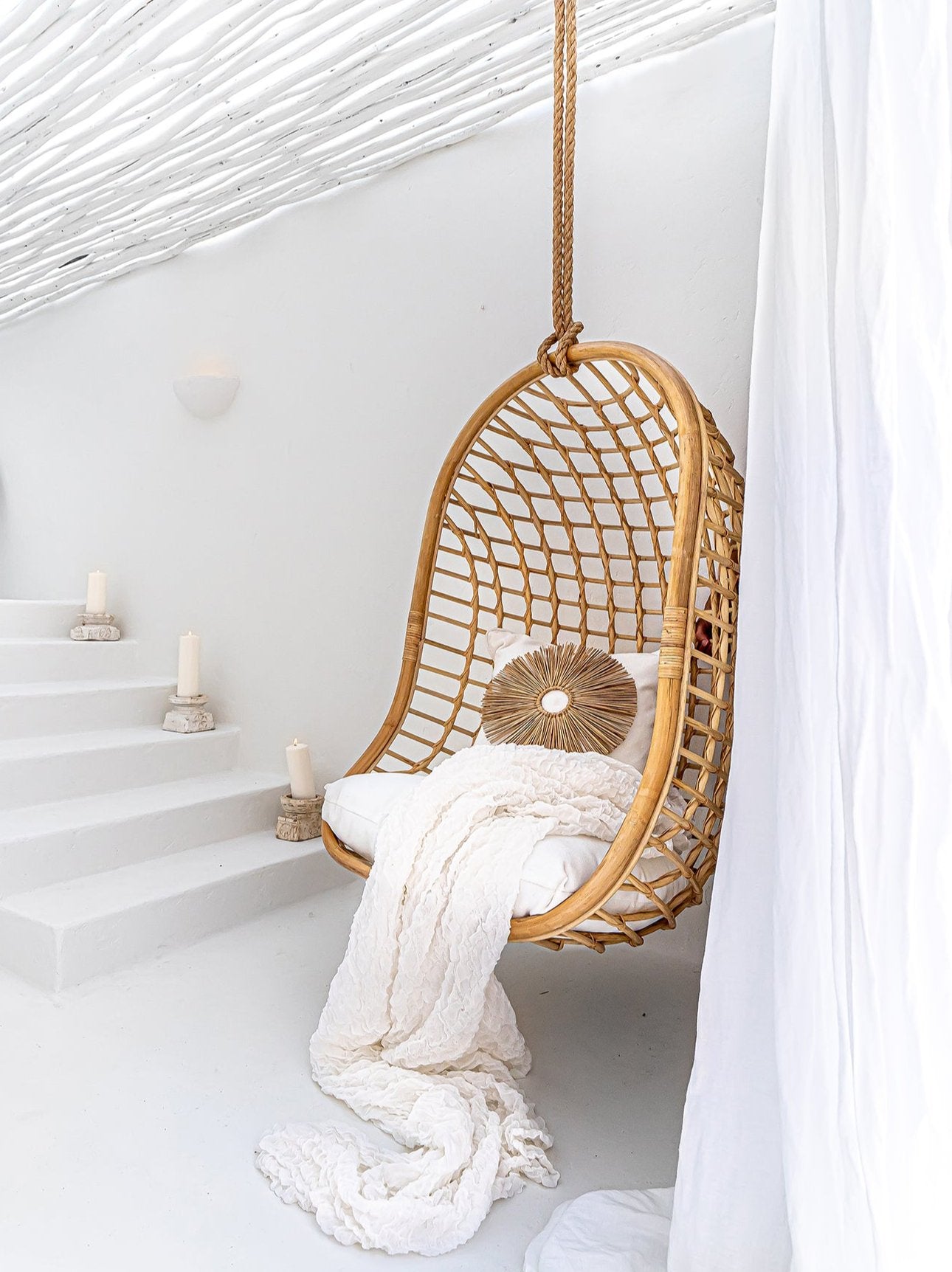 A natural rattan hanging chair with an open-weave design, suspended by a rope in a serene, all-white setting. Styled with plush white cushions, a textured throw, and a decorative sunburst pillow, creating a tranquil and inviting retreat