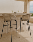 Natural rope outdoor bar stools with curved backs and black metal legs styled at a textured stone bar on a coastal balcony with margaritas and snacks