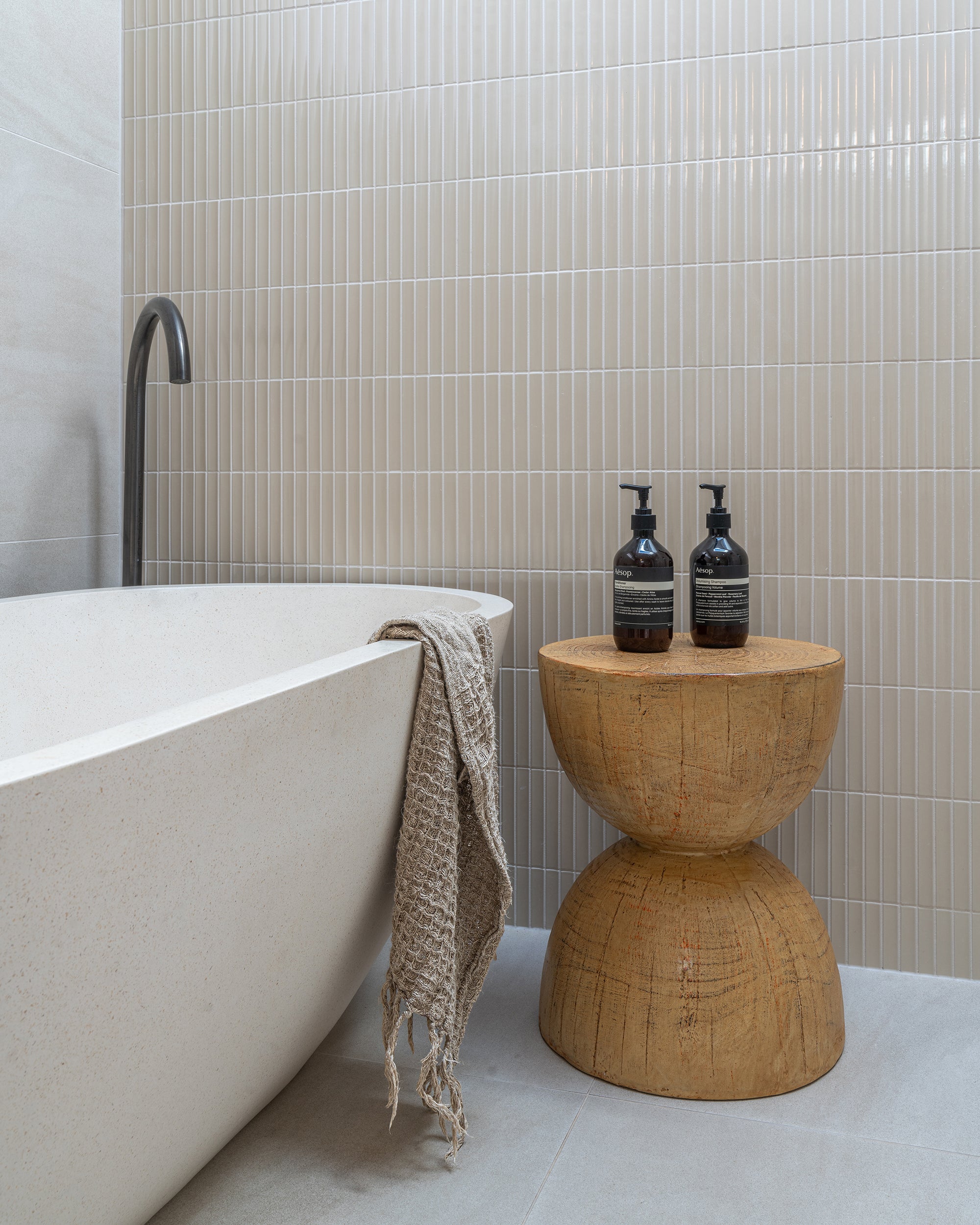 Modern bathroom featuring a freestanding stone bath, black tapware, and a carved timber hourglass side table with Aesop bottles and a neutral-toned towel.