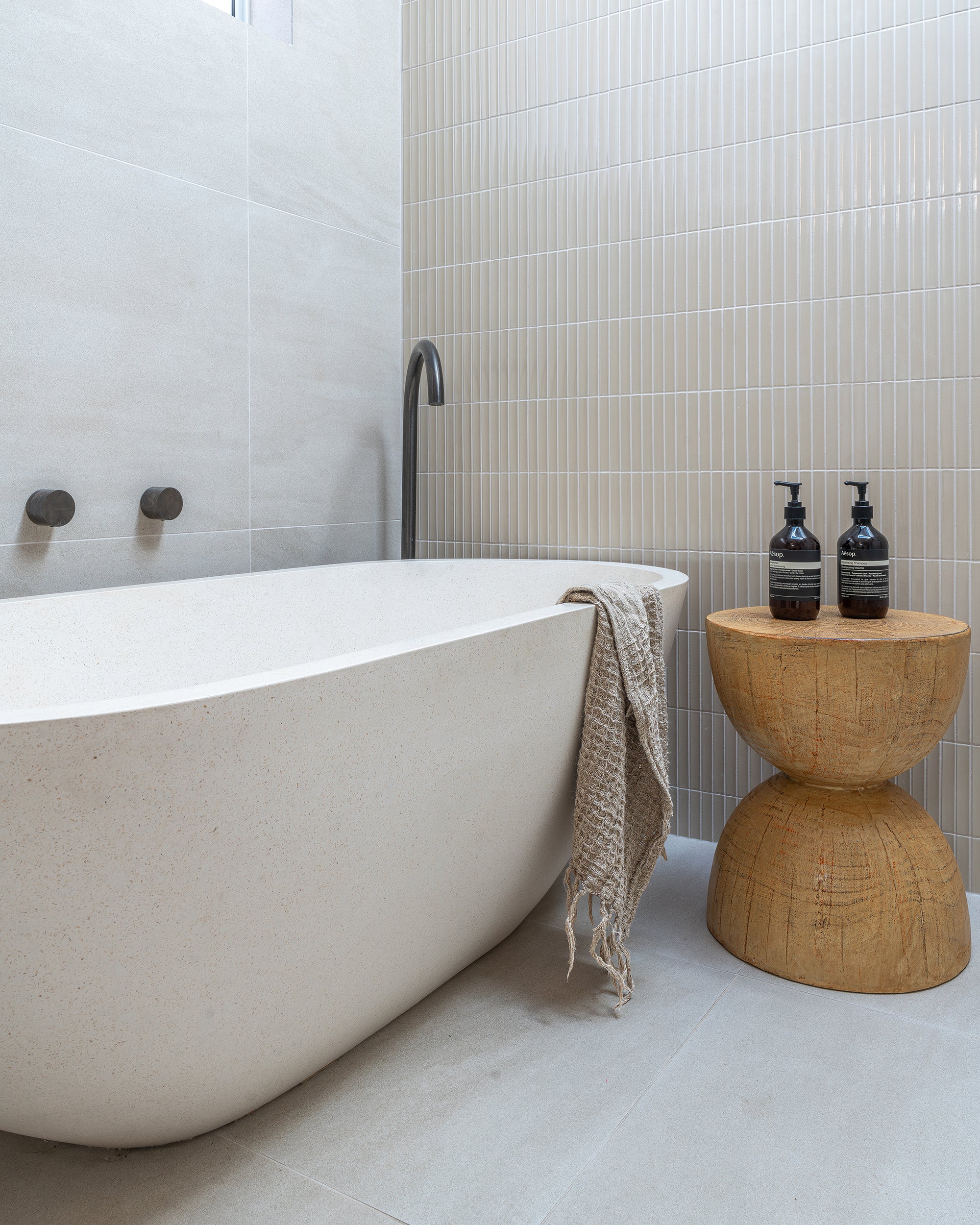 Modern bathroom featuring a freestanding stone bath, black tapware, and a carved magnesium oxide hourglass side table with Aesop bottles and a neutral-toned towel.