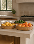 Three woven natural fibre trays filled with lemons, limes, and oranges styled on a rendered kitchen bench in a Mediterranean-inspired space with soft filtered light.
