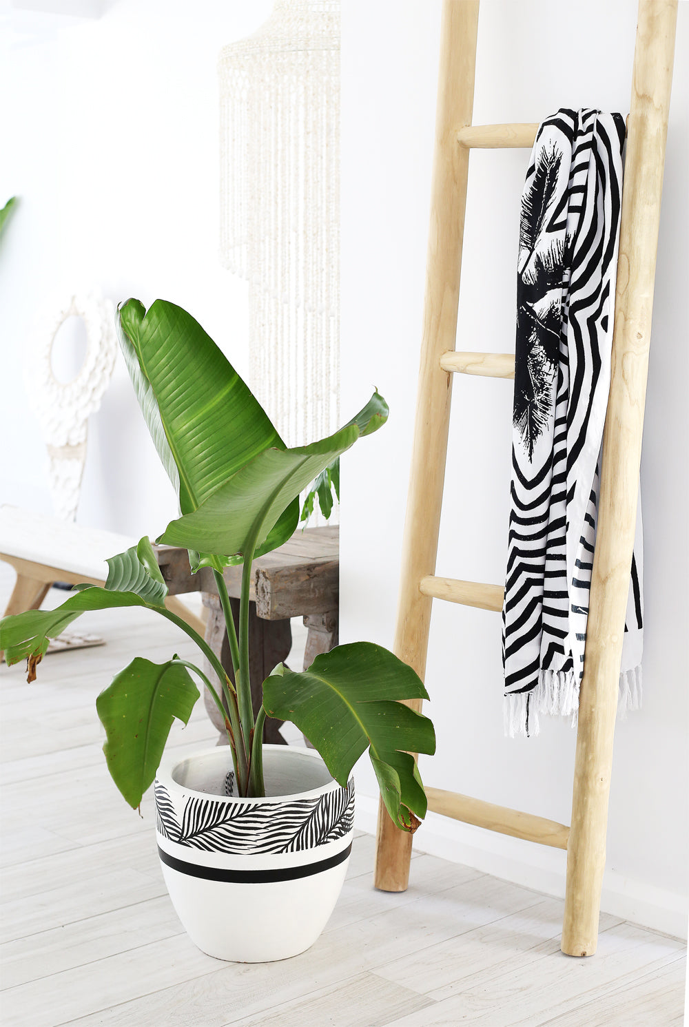 A natural wooden ladder styled as a towel and blanket rack in a modern boho-inspired interior. Paired with a tropical plant in a black-and-white patterned pot, this piece adds warmth and texture to any home