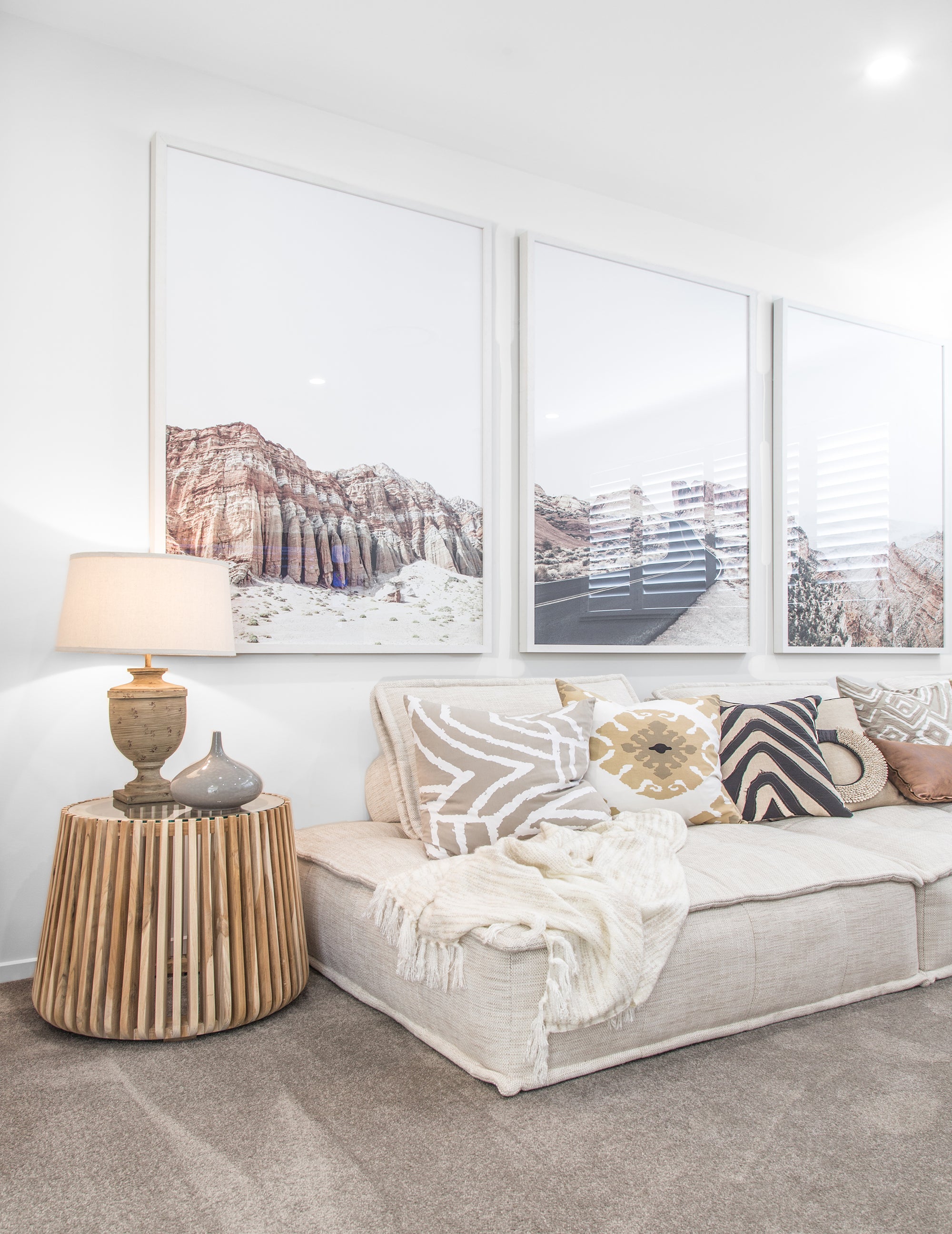 A contemporary lounge space featuring a low-profile neutral sofa adorned with patterned and textured cushions, a wooden slatted side table, and a ceramic table lamp. Large framed desert landscape artwork adds a serene, earthy aesthetic to the room
