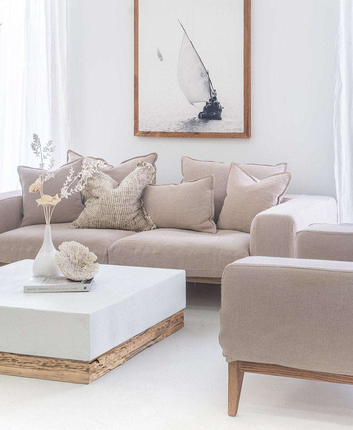 A serene and stylish living room featuring a neutral-toned sofa adorned with textured cushions, a white and wood coffee table, and a framed black-and-white sailing artwork. The light-filled space exudes a calming coastal aesthetic with natural textures and soft, earthy hues