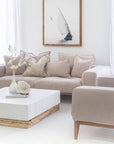 A serene and stylish living room featuring a neutral-toned sofa adorned with textured cushions, a white and wood coffee table, and a framed black-and-white sailing artwork. The light-filled space exudes a calming coastal aesthetic with natural textures and soft, earthy hues