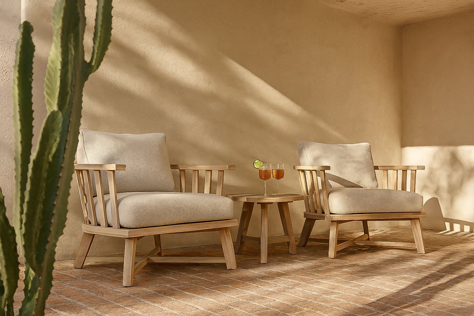 Nikki Timber Occasional Chairs with teak frames and natural upholstered cushions styled in an outdoor lounge with a round timber side table and warm afternoon light