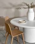 A contemporary dining space featuring Kulala Living oak wood dining chairs paired with a white round table with a fluted base. The setting is styled with a textured ceramic vase, dried foliage, and neutral-toned dinnerware, creating a serene and modern aesthetic