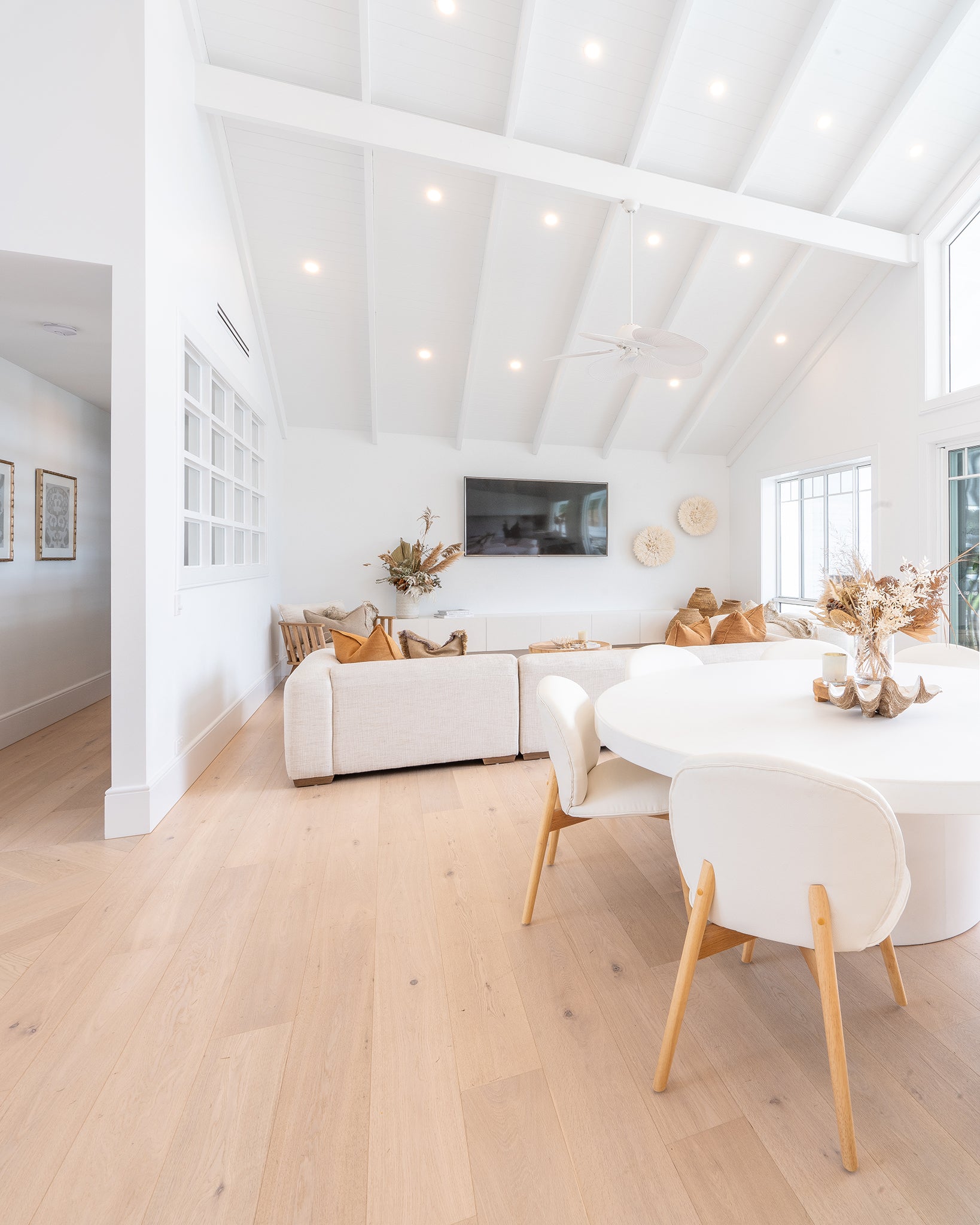 Open-plan lounge and dining room with vaulted white ceiling and natural decor elements, featuring a neutral modular sofa, round white dining table with upholstered chairs, dried floral arrangements, and wooden accents.